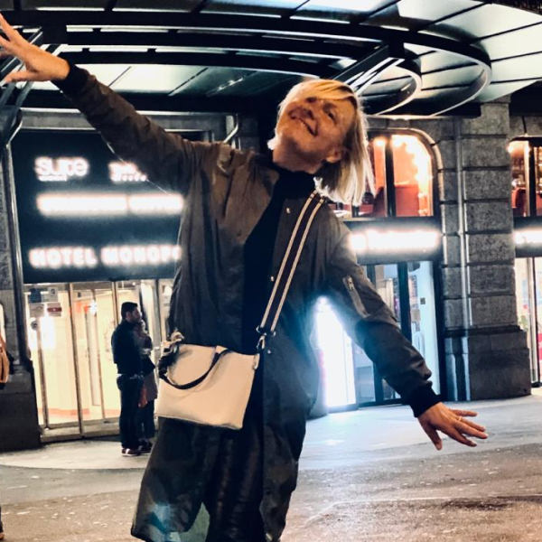 A woman with short blonde hair smiling and posing with arms outstretched outside a building at night, illuminated by streetlights.