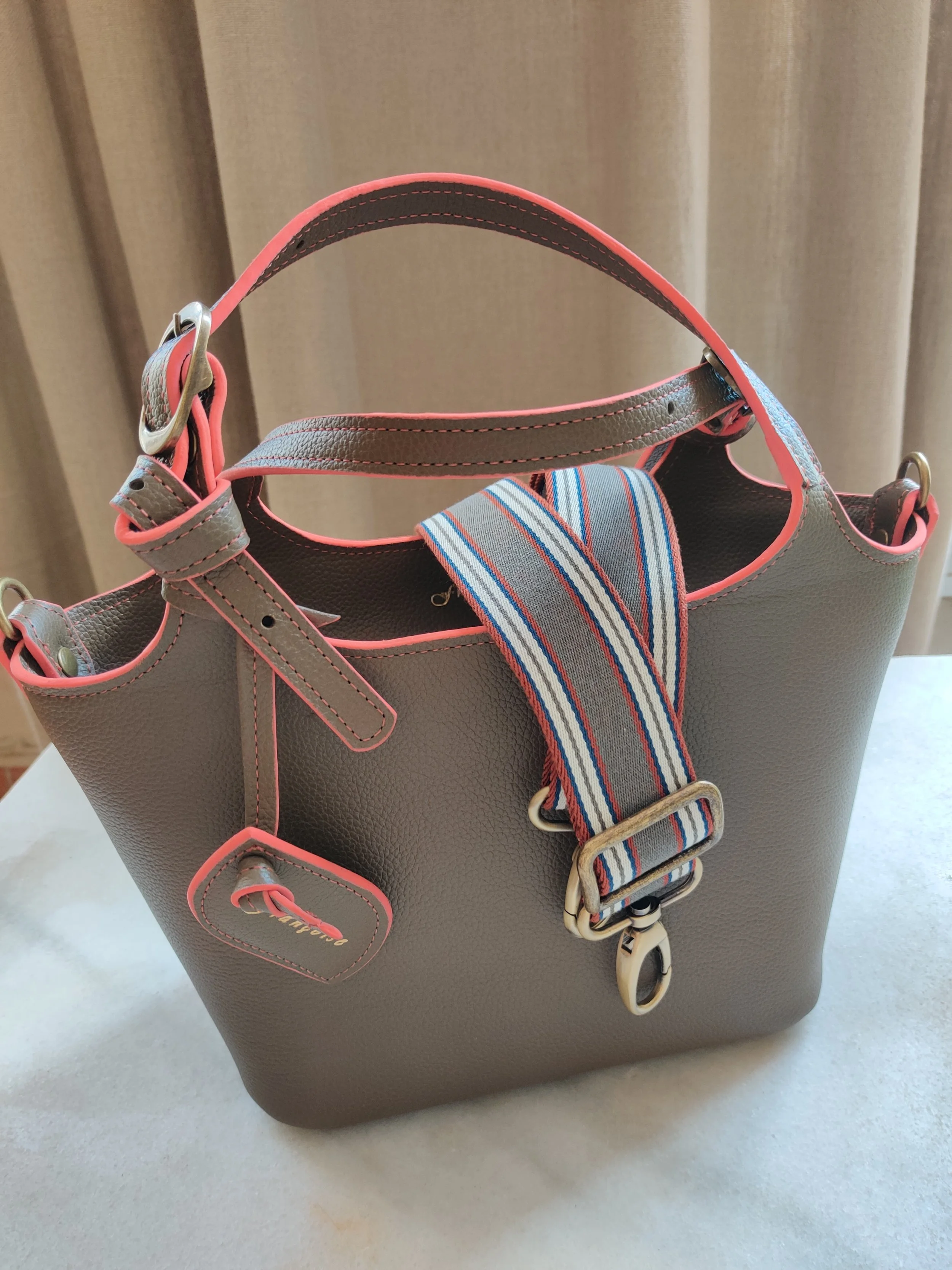 A grey handbag with grey straps with a pink trim, placed on a white surface, featuring a striped fabric strap and a small matching tag attached.