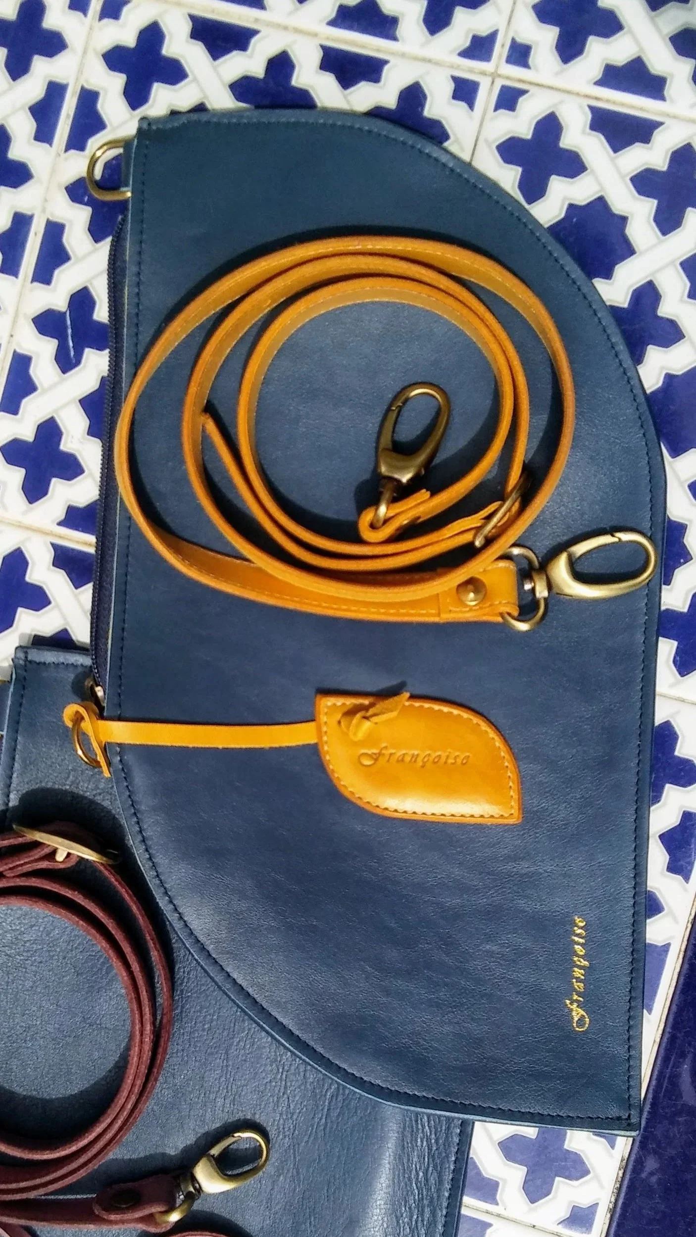 A navy Françoise Cross Body leather handbag with contrasting yellow leather detachable leather strap, placed on a blue and white tiled surface.
