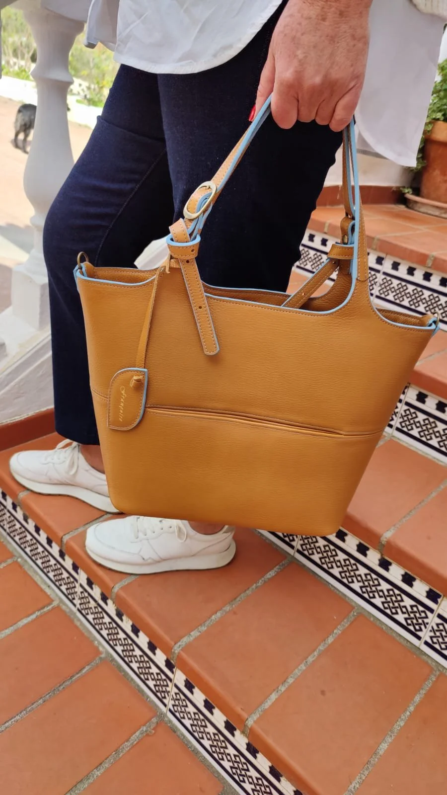 A person holding a Francoise Choco Late leather handbag with blue accents, standing on terracotta tiled stairs with decorative black and white tiles, wearing white sneakers and dark blue jeans.