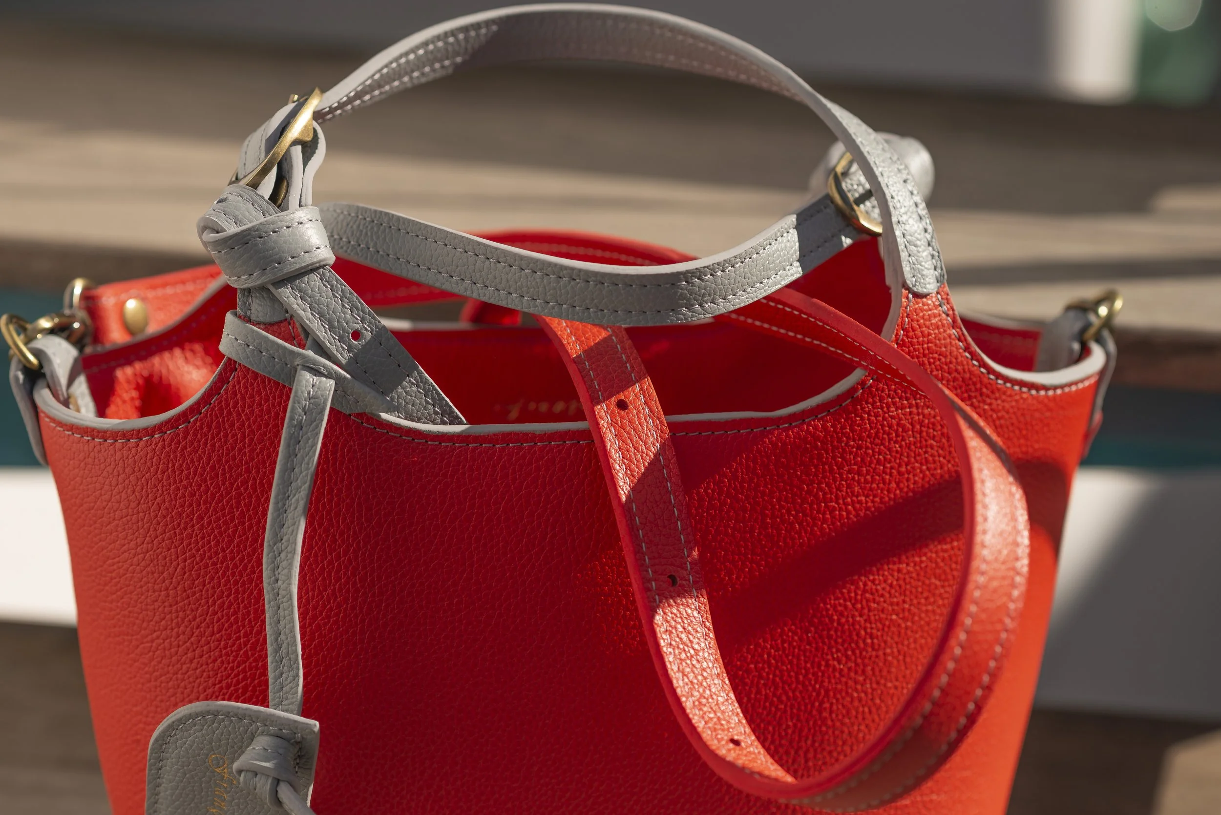 A red Francoise Noisette leather handbag with gray and red straps, placed on a wooden surface in sunlight.