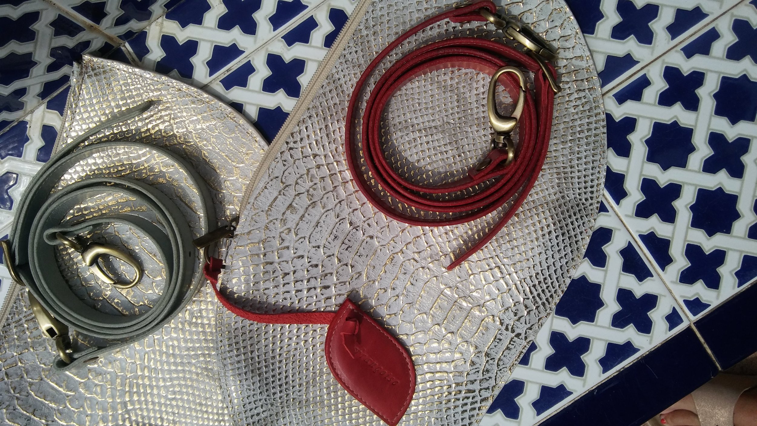 A silver textured Françoise Crossbody leather handbag with a red rolled-up belt. The background includes blue and white tiles. 