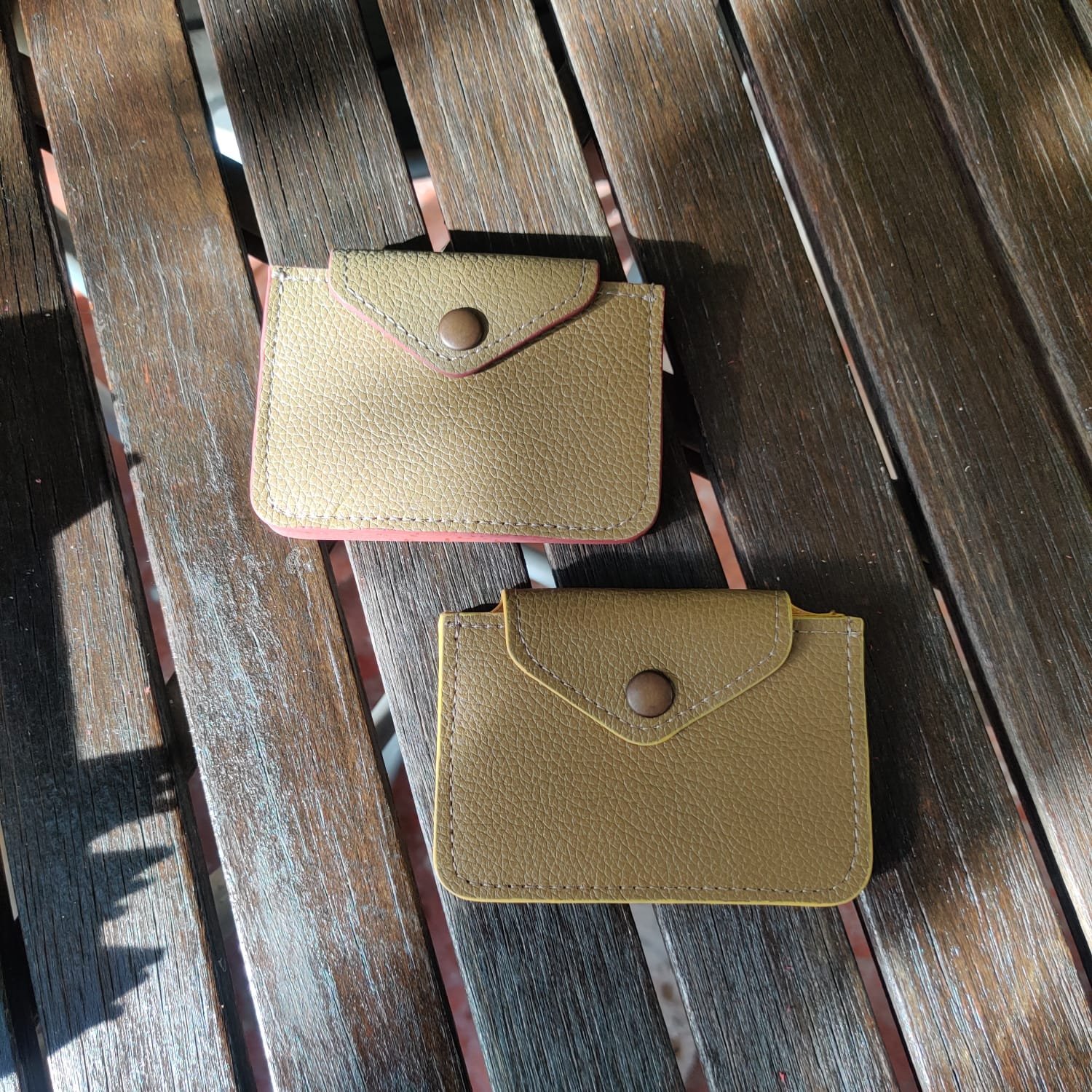 Two small cardholders with flaps and snap buttons are placed on a wooden surface outdoors. One wallet is beige with pink edges, and the other is beige with yellow edges.