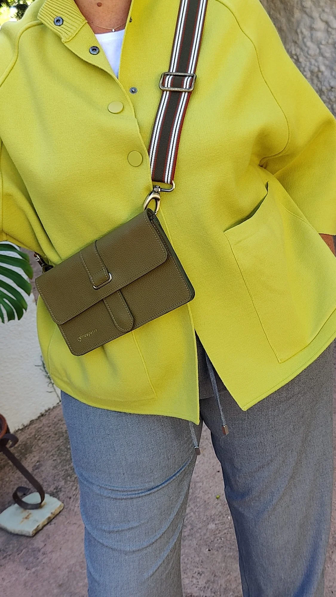 Person wearing a bright yellow jacket carrying an dark green Françoise City leather handbag with a striped canvas shoulder belt, standing outdoors near a wall and plant.