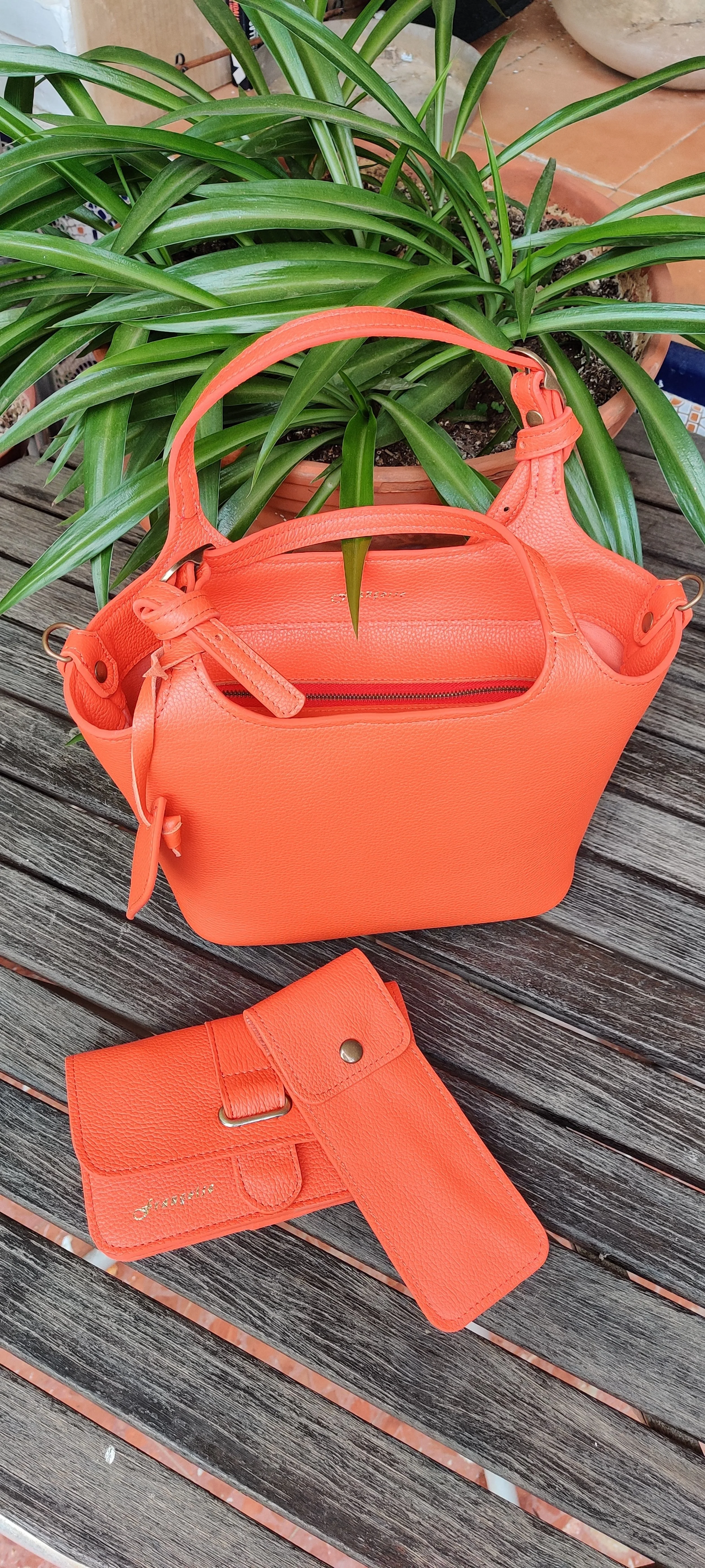 An orange Francoise Noisette leather handbag with matching wallet and pencil case holder placed on a wooden outdoor table beside a potted green plant.