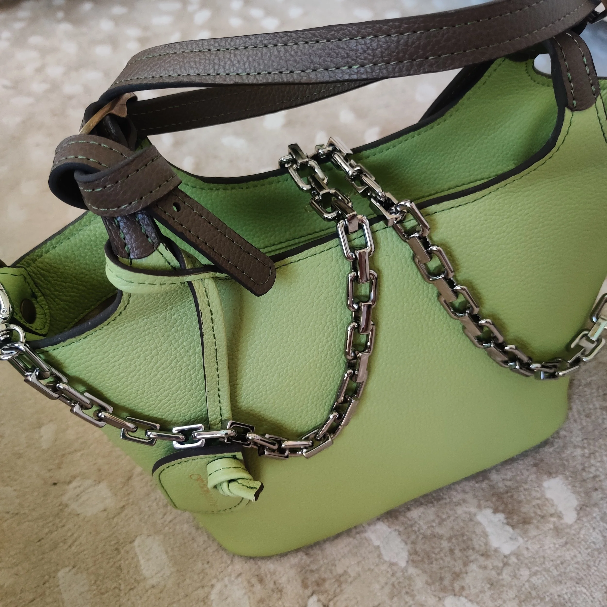 A lime green handbag with a dark brown leather handle and silver belt, placed on a beige patterned surface.