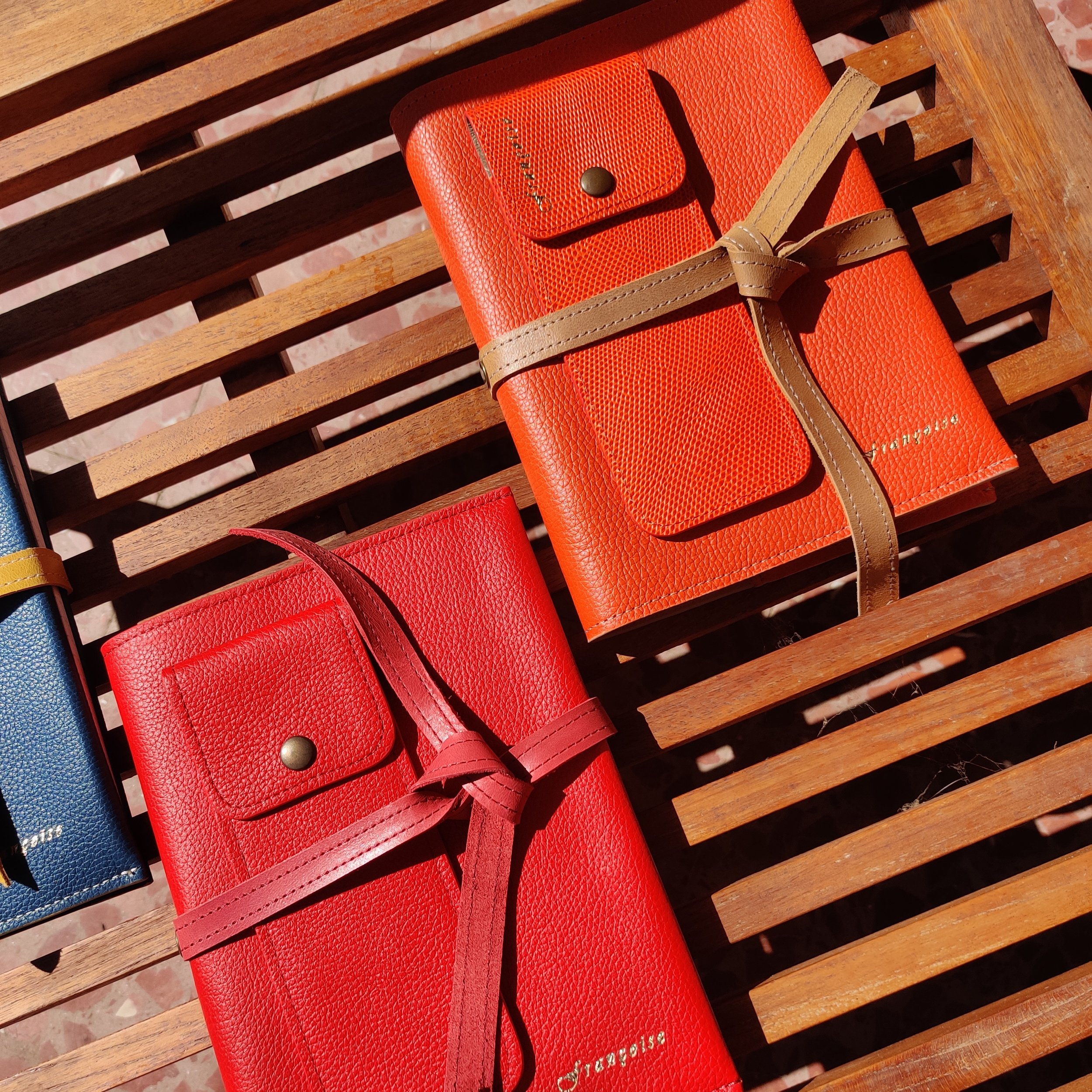 Three colourful leather notebooks are placed on a wooden slatted surface outdoors. The notebooks are red, orange, and blue, with gold and black accents.