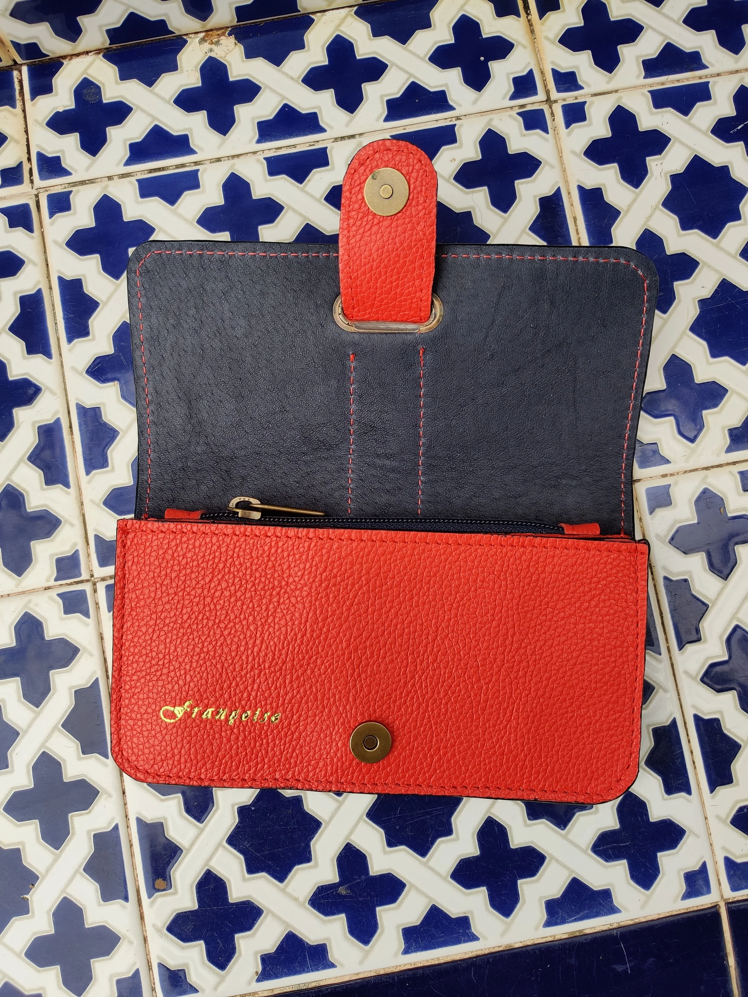 A red leather wallet with a dark blue inside, with snap closure, on a blue and white patterned tile floor.