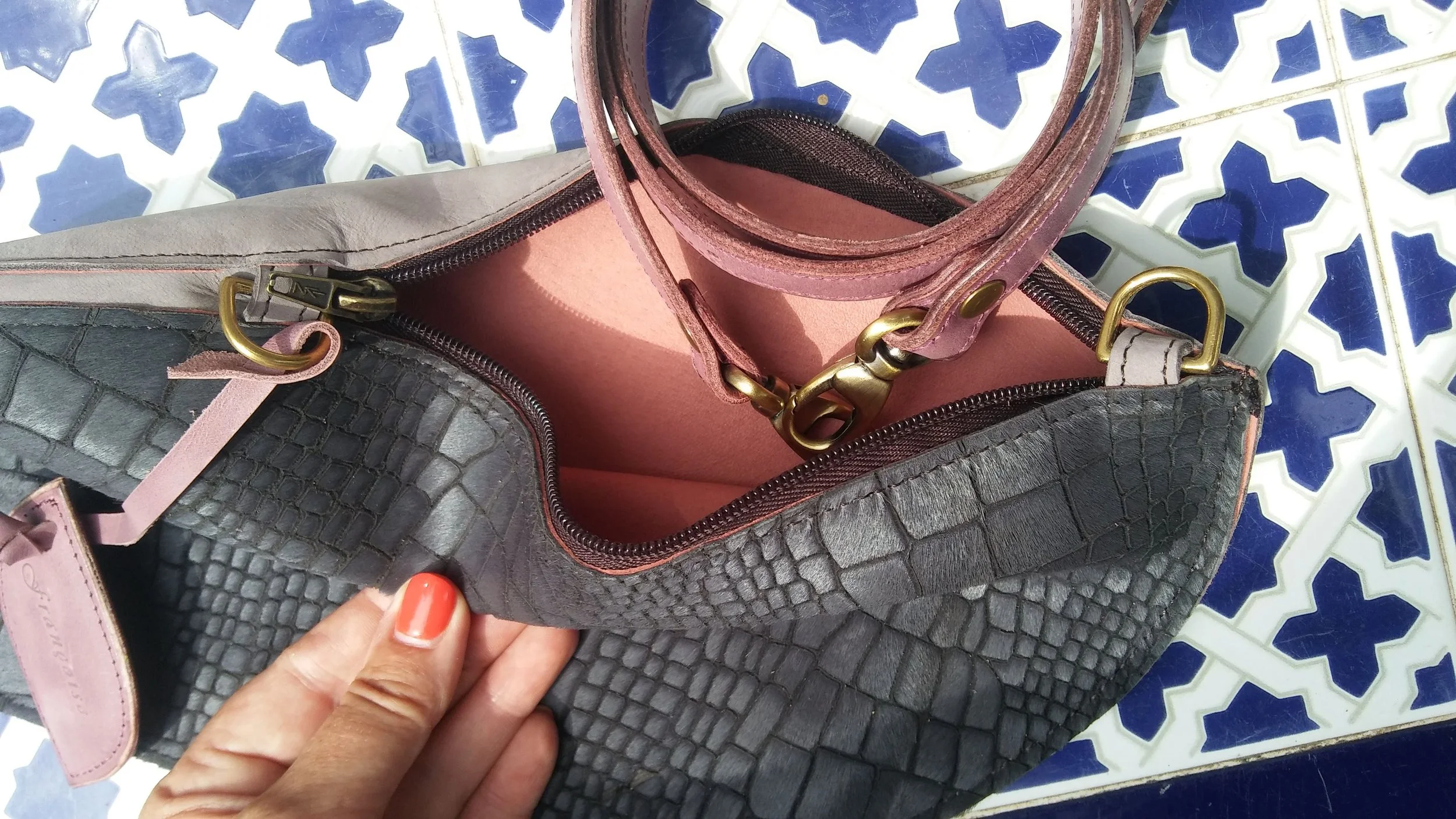 Close-up of a Françoise Cross Body leather handbag with gray crocodile texture, partially open, on a blue and white patterned tile surface. Inside the bag, there is a pink inner lining and a matching pink strap. 