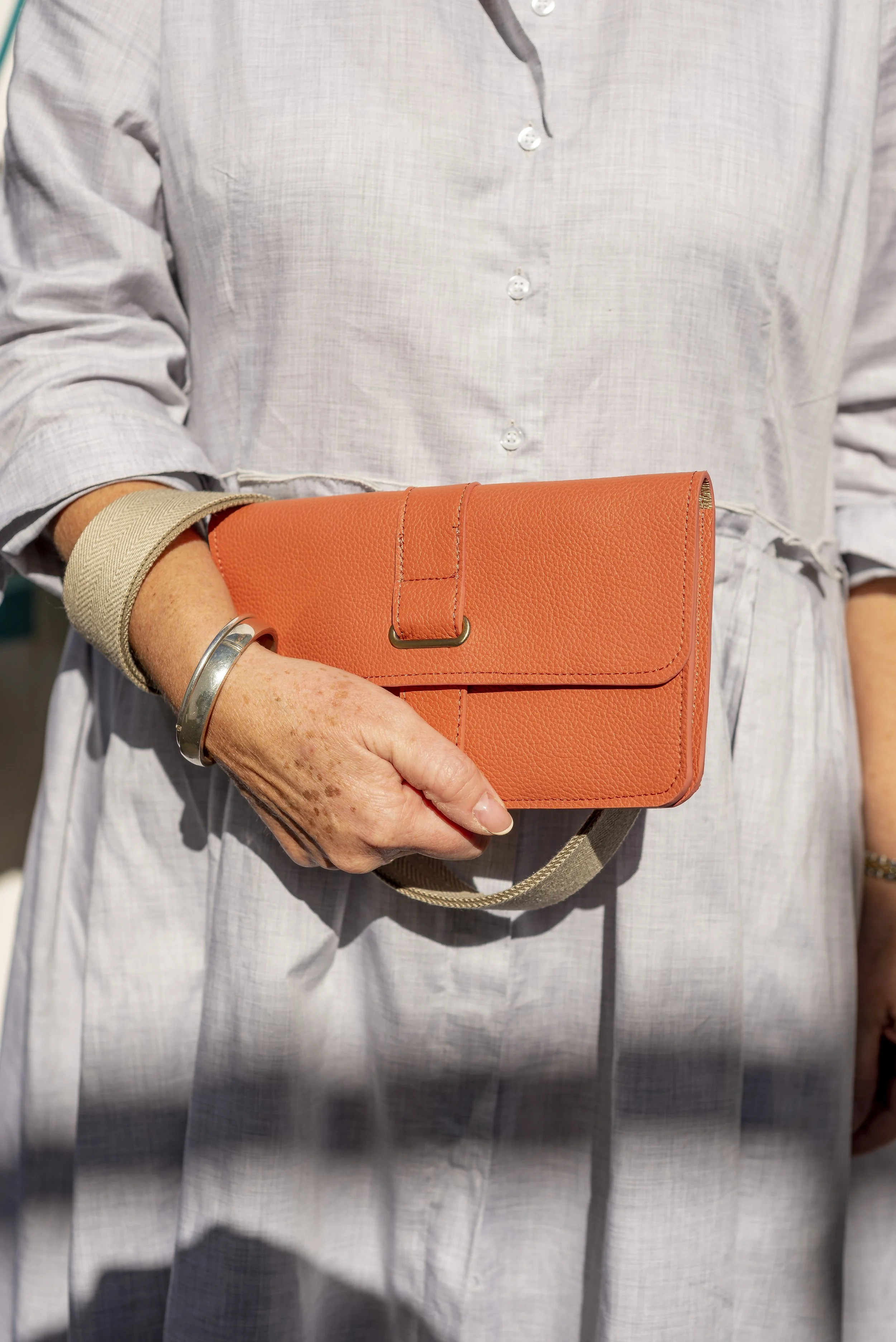 Person holding an orange Françoise City leather handbag, wearing a light gray button-up linen dress with rolled-up sleeves and silver bracelet.