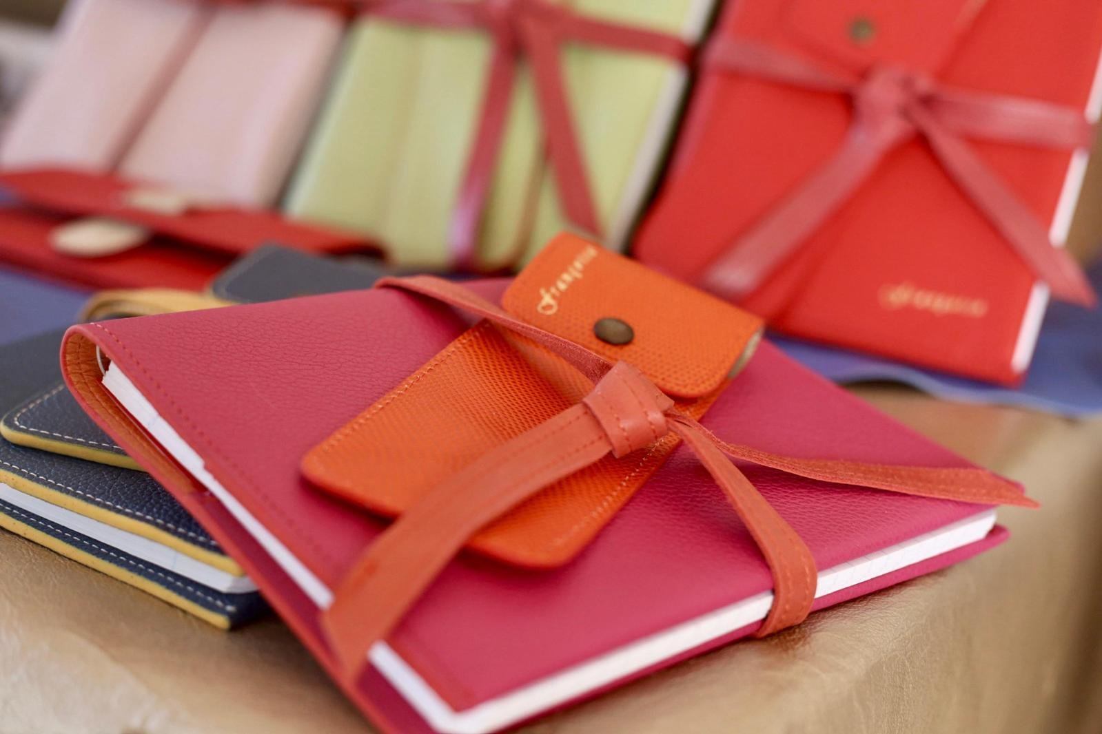 A pink notebook with an orange pencil case, placed on top of a pile of other notebooks. In the background, there are more notebooks with ribbons.