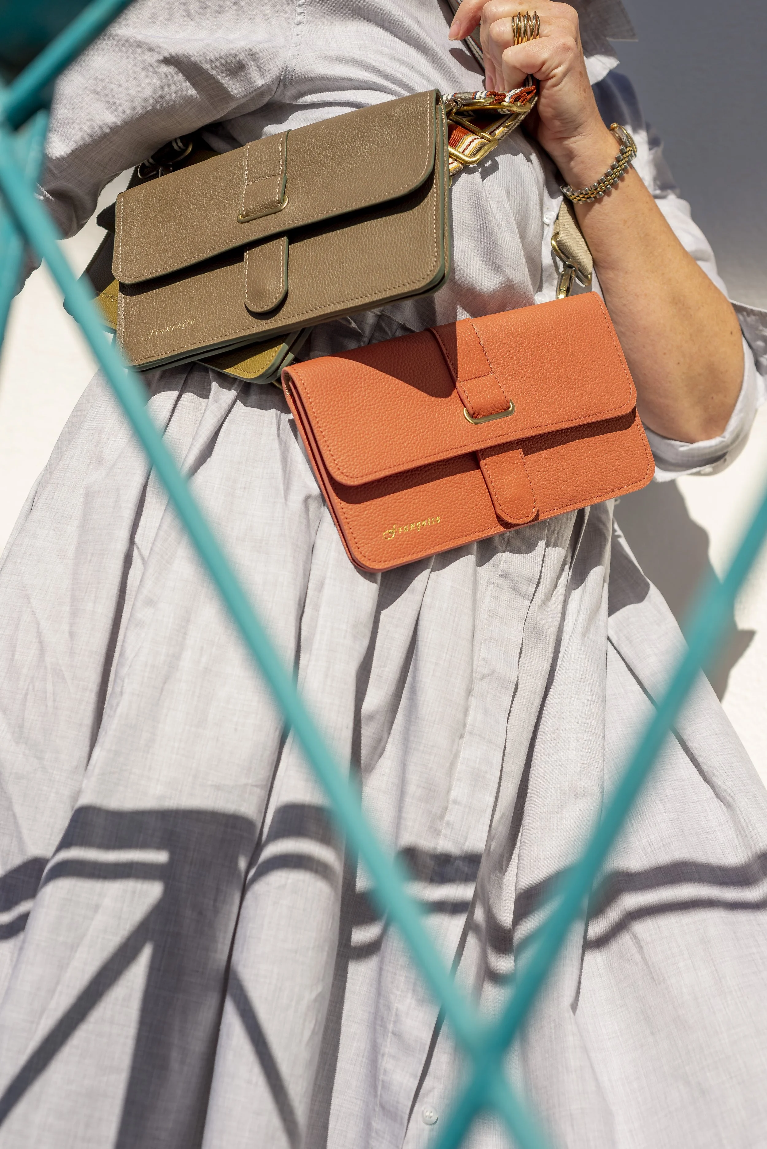A person wearing a white dress with two Françoise City leather handbag, one tan and the other orange, over their shoulder. The image is taken through a blue metal fence.