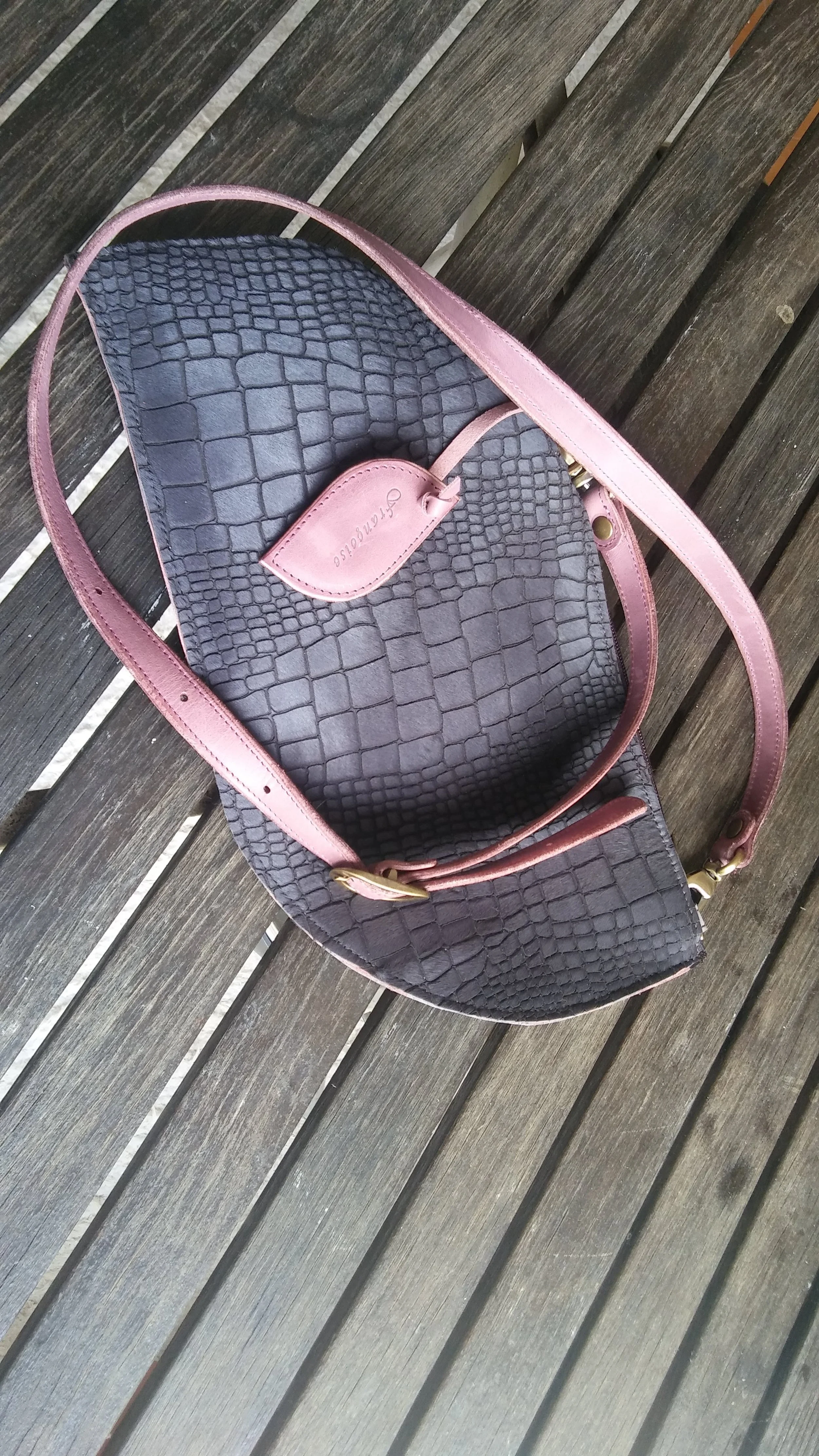 A Françoise Cross Body leather handbag with a snake skin resembling texture, featuring a pink leather straps and a pink tag, resting on a wooden surface.