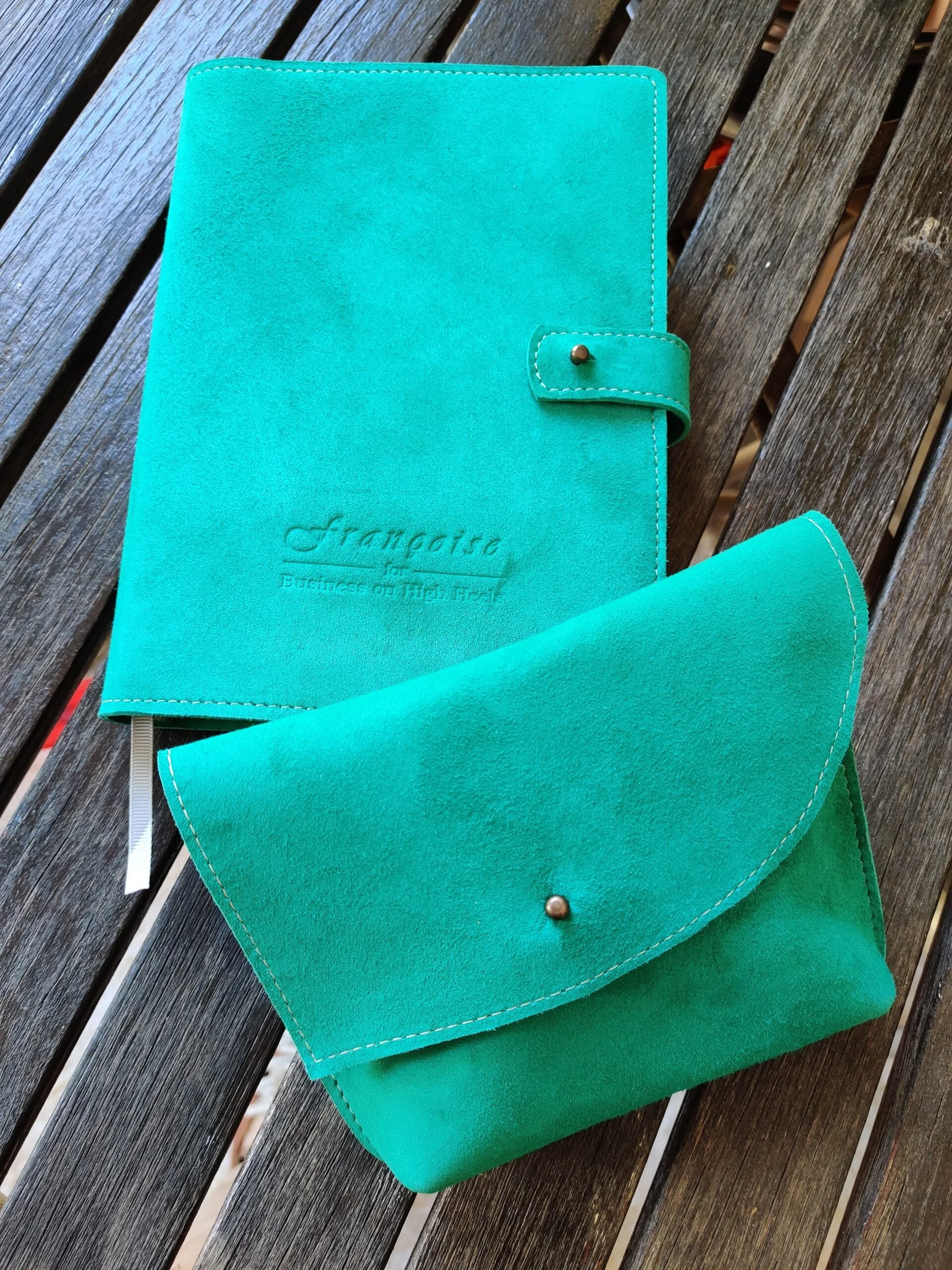 A turquoise suede planner with embossed text, a matching pen, and a small clutch or wallet on a wooden surface.