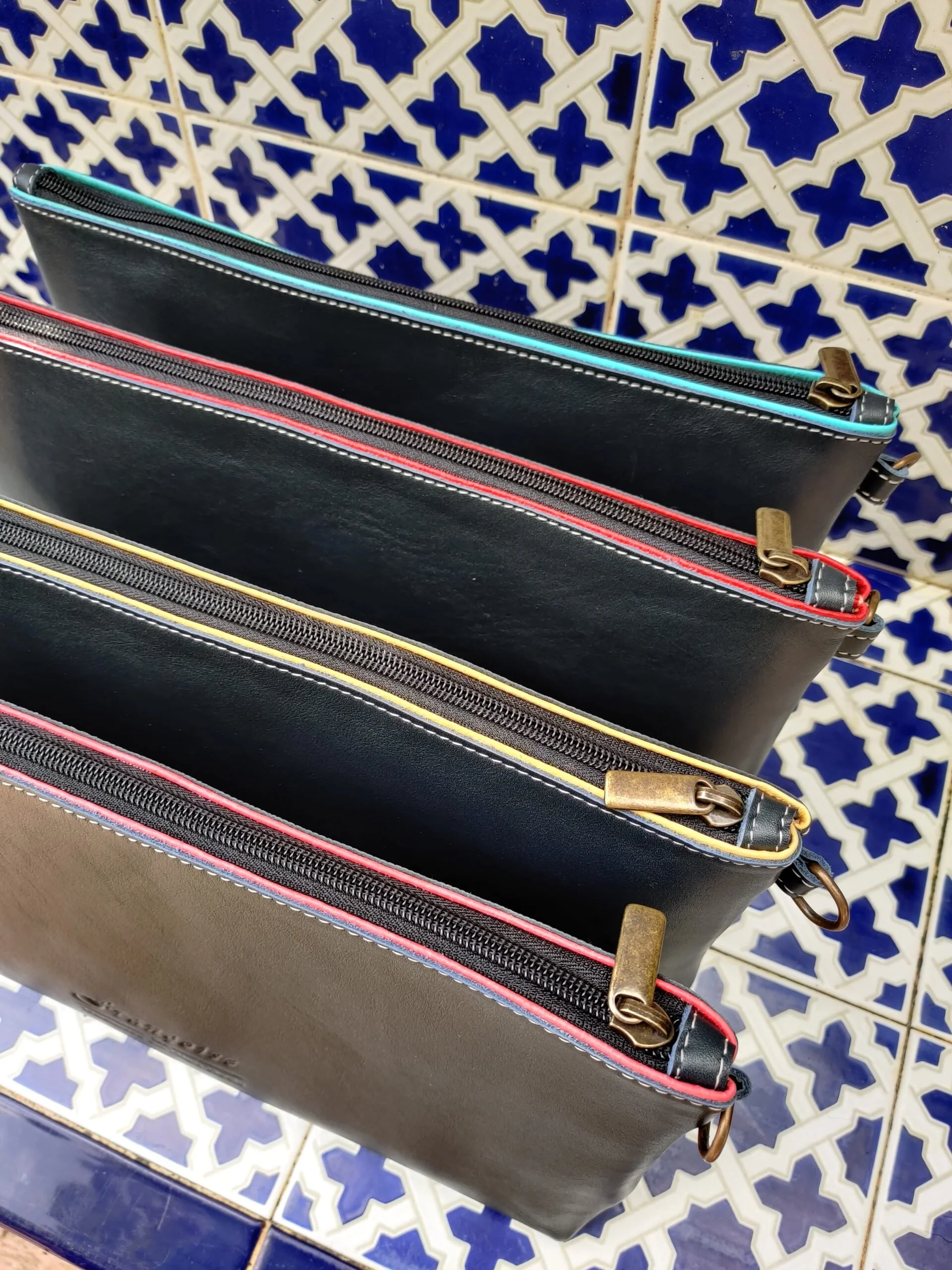 Four leather zippered cases with colourful zips against a blue and white patterned tile background.