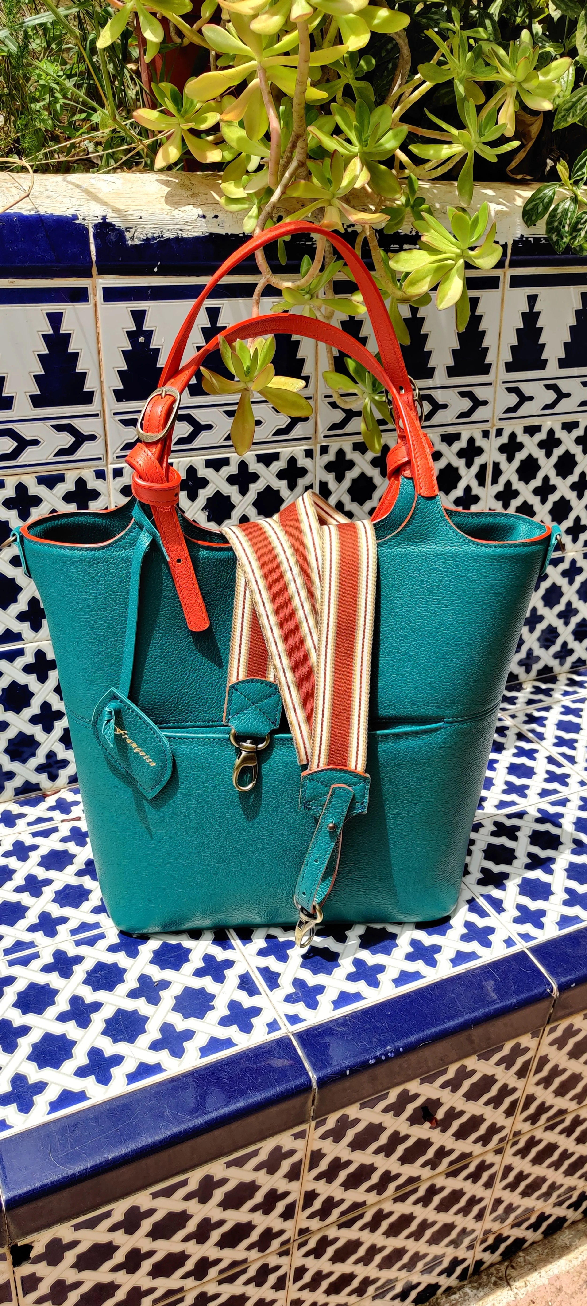 A teal Francoise Caramel leather handbag with orange handles, placed on a decorative blue and white tiled surface, with green leafy plants in the background and a canvas belt hanging from the bag.