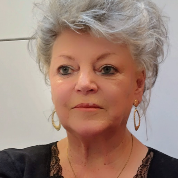 A woman with short, curly gray hair wearing gold hoop earrings and a black top with lace details, standing indoors against a plain wall.