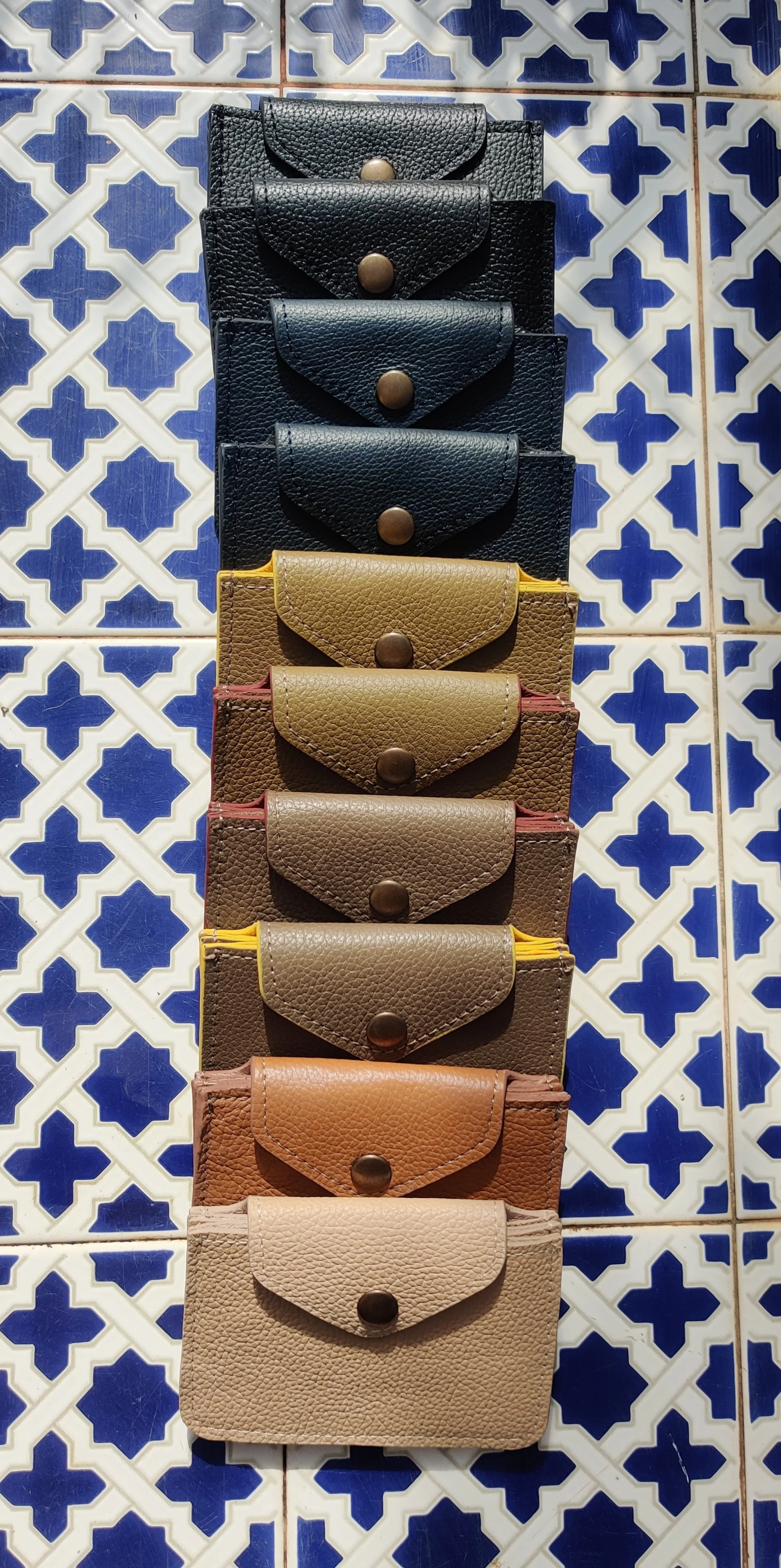 A vertical display of leather cardholders in different colours, ranging from black, dark brown, yellow, to beige. The cardholders are placed on a blue and white geometric tiled background.