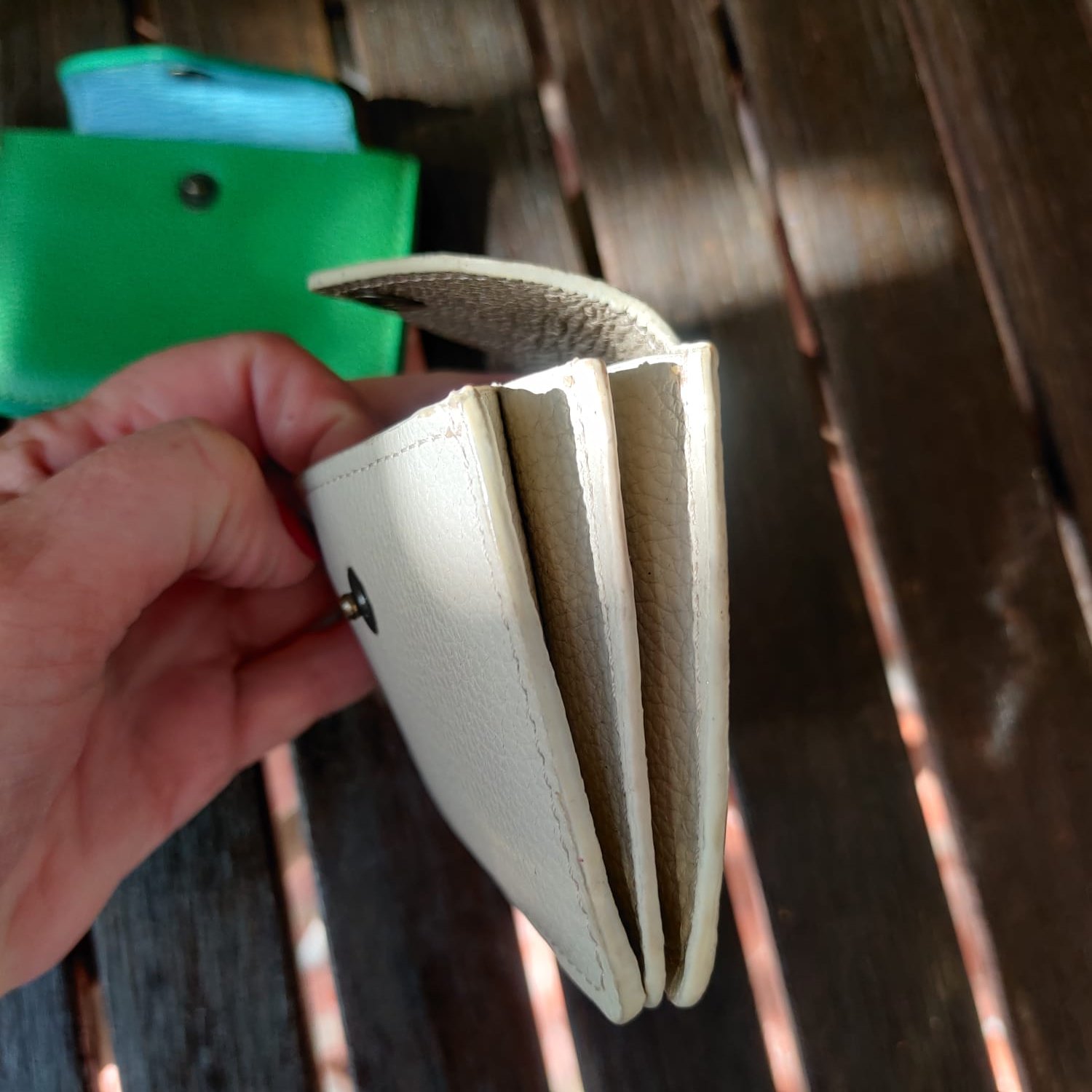 A person's hand holding an open cardholder with visible compartments, on a wooden surface with a green cardholder in the background.
