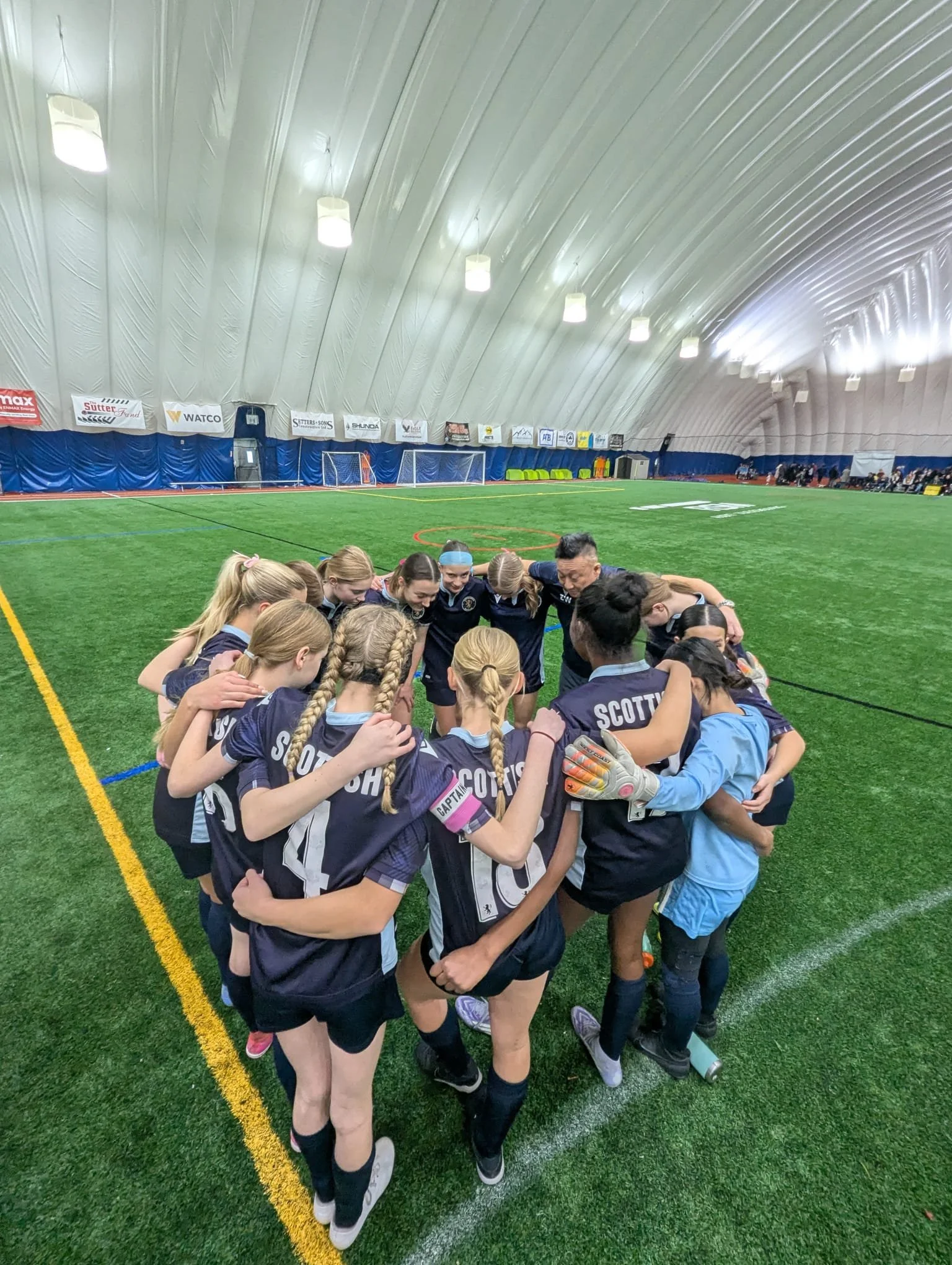 A youth athletics sports soccer team huddles in a circle on an indoor field, with their coach leading an inspirational and motivational pre game speech.