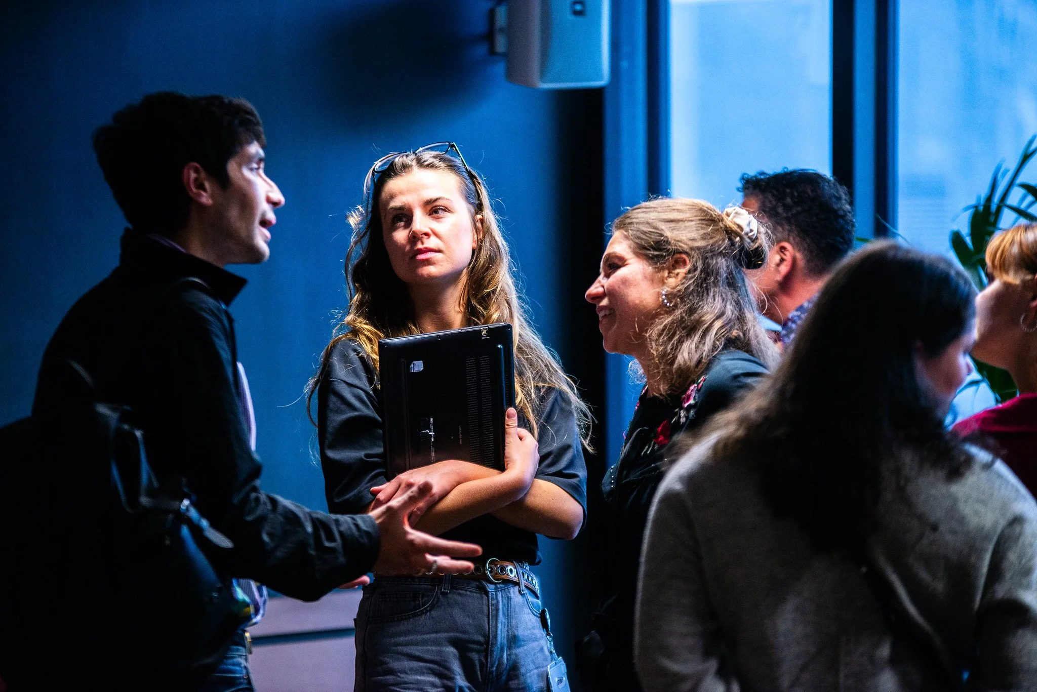 Group of people having conversations in an indoor setting, two women in the center engaged in a discussion, one holding a laptop, with a blue wall and window in the background.