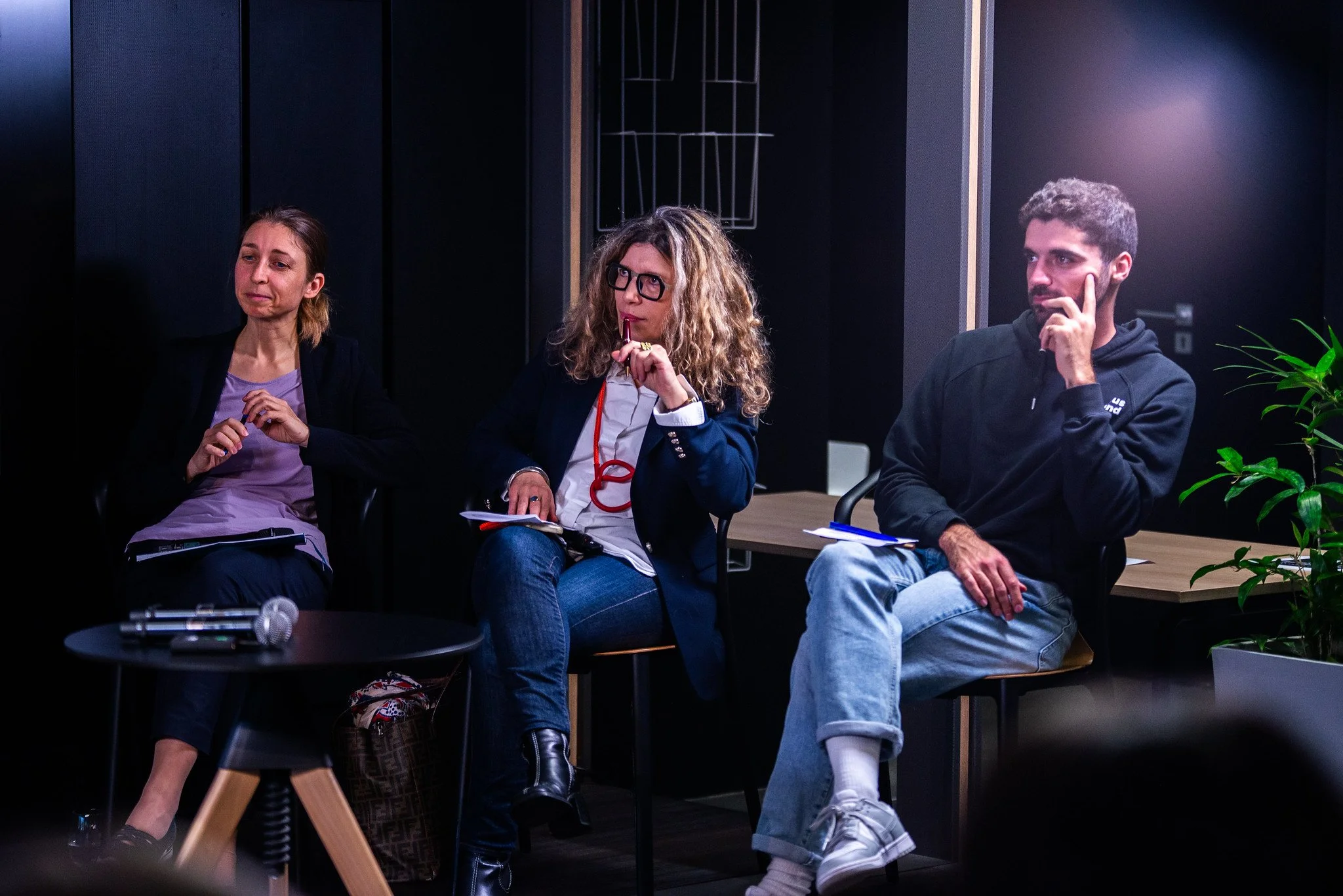 Three people sitting in a room, seemingly during a discussion or workshop. The woman on the left has her eyes closed and appears to be emotional. The woman in the middle has curly hair, glasses, and is holding a pen, with a thoughtful expression. The