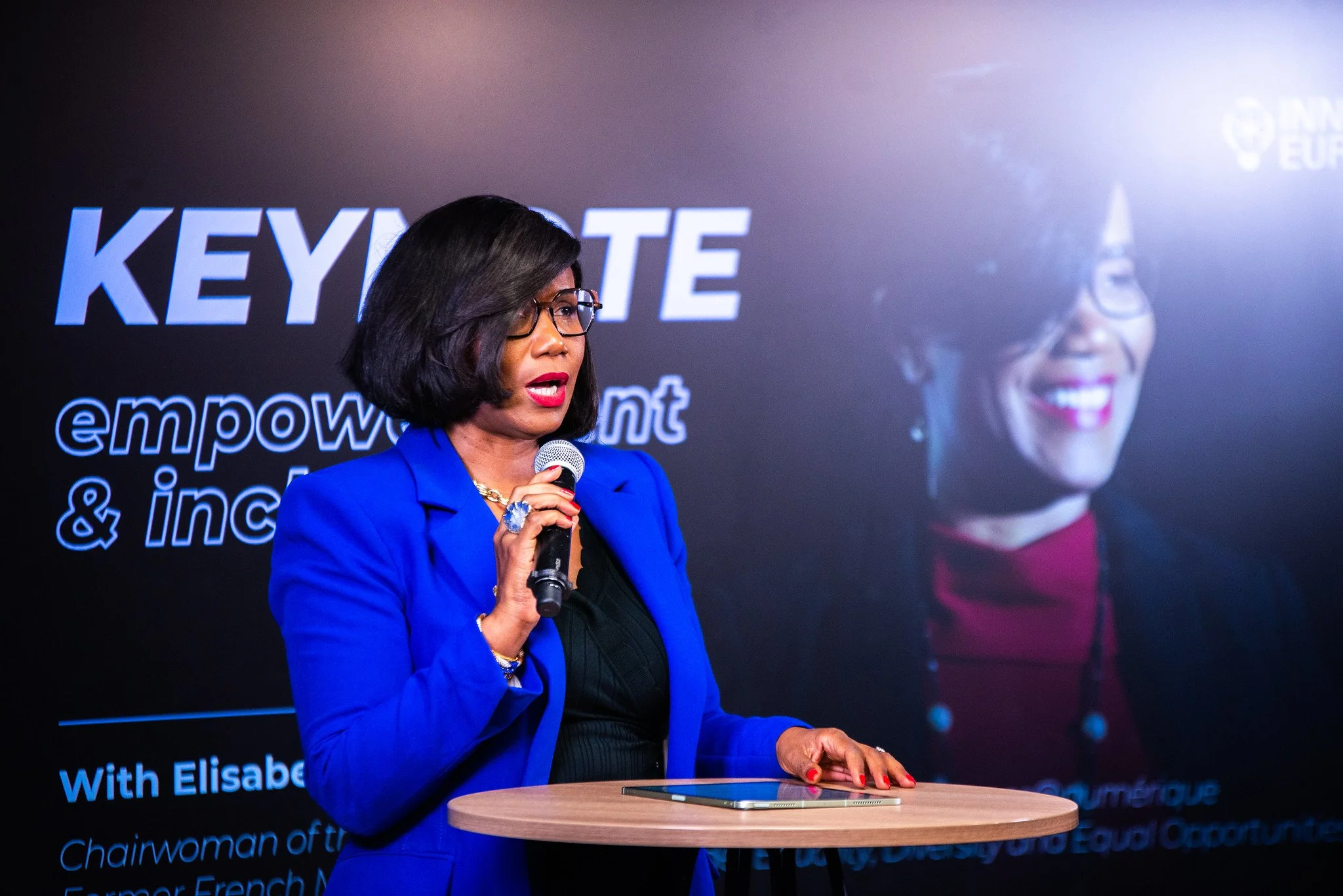 A woman in a blue blazer speaking into a microphone at a presentation or conference, with a large screen behind her displaying the text 'KEYNOTE' and her image.