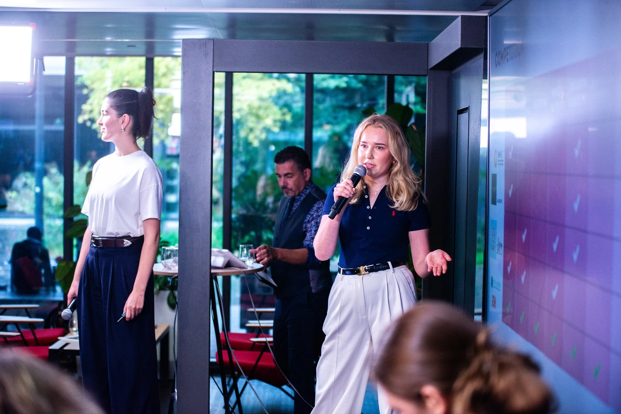 Teenage girl speaking into a microphone at an indoor event. She has blonde hair and is wearing a navy polo shirt and white pants. Nearby, a woman in a white top and navy pants stands with a microphone in her hand, and a man in a vest works on a lapto