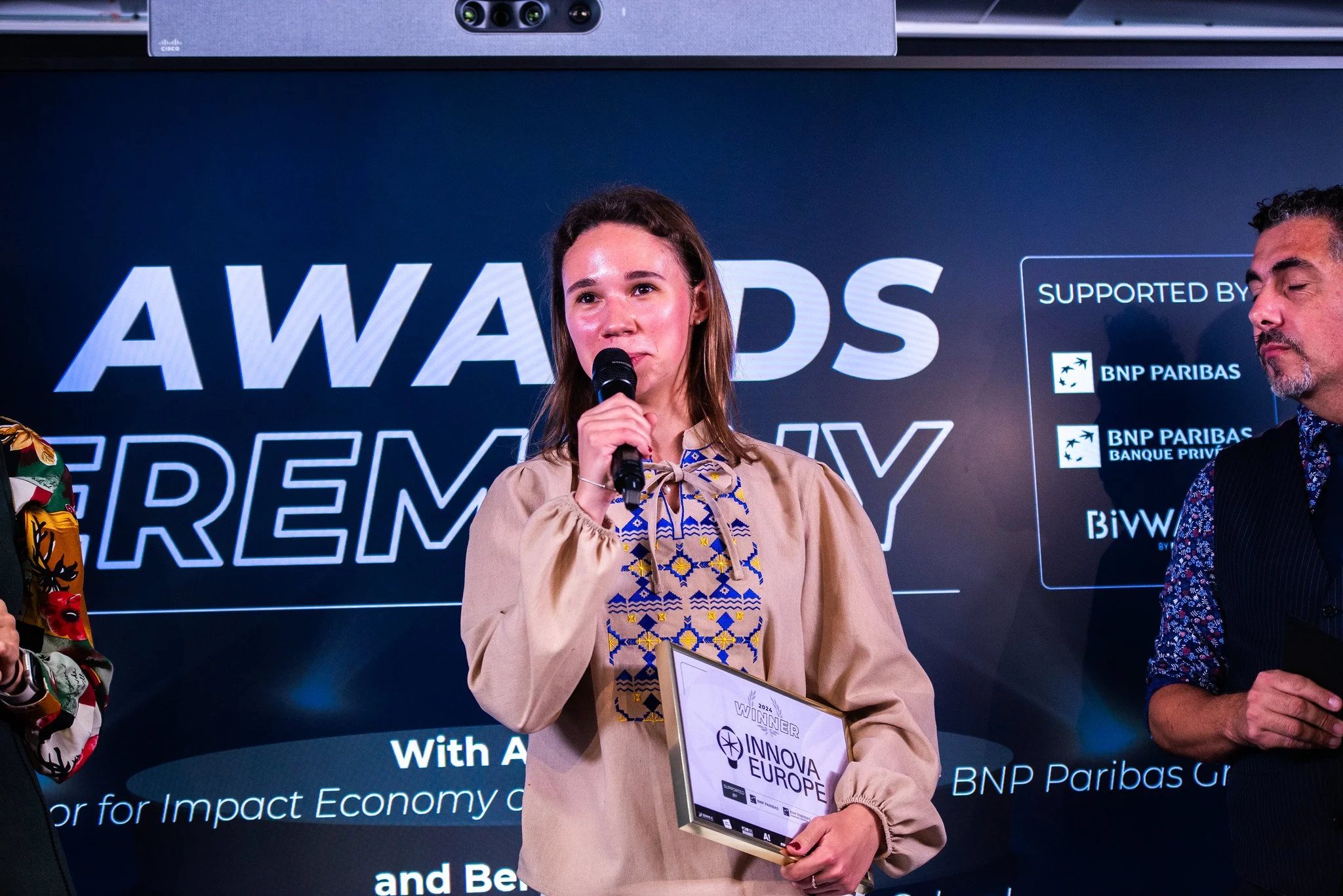 Young woman in traditional embroidered blouse holding a framed award, speaking into a microphone at an awards ceremony with a large screen behind her showing the words "Awards Ceremony" and logos of supporting companies.