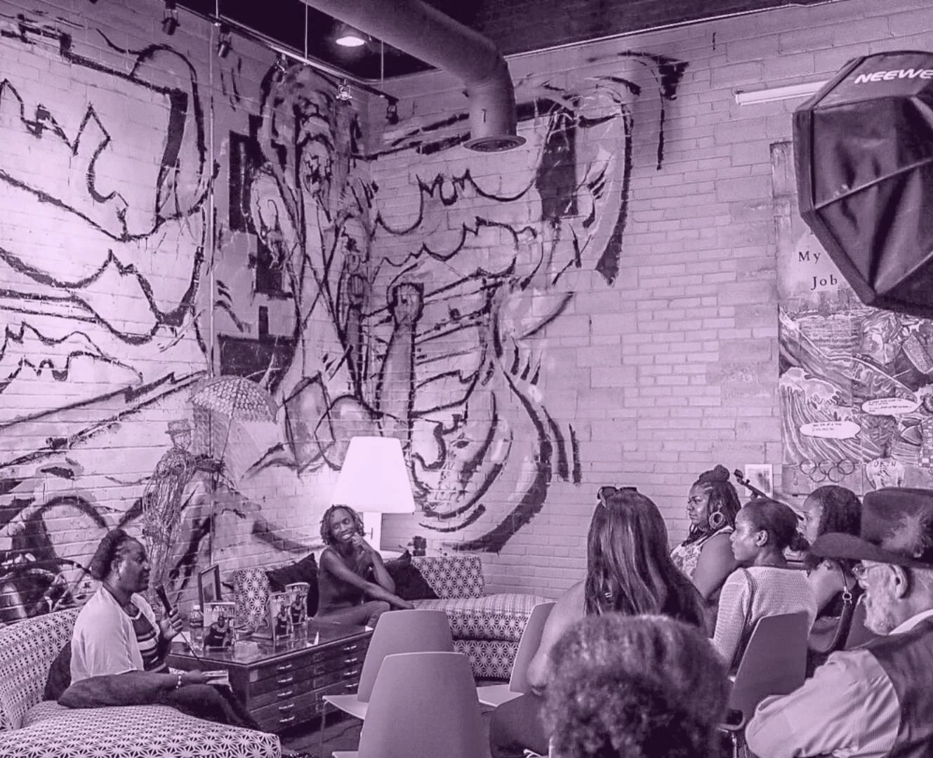 People seated at a Sit With Me event, with a speaker presenting. The room has a large artwork or mural on the brick wall behind them, showing abstract or expressive faces and figures.