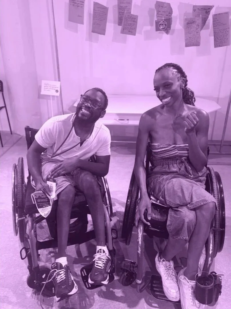 Two people in wheelchairs smiling and looking happy, inside a room with an art installation behind them.