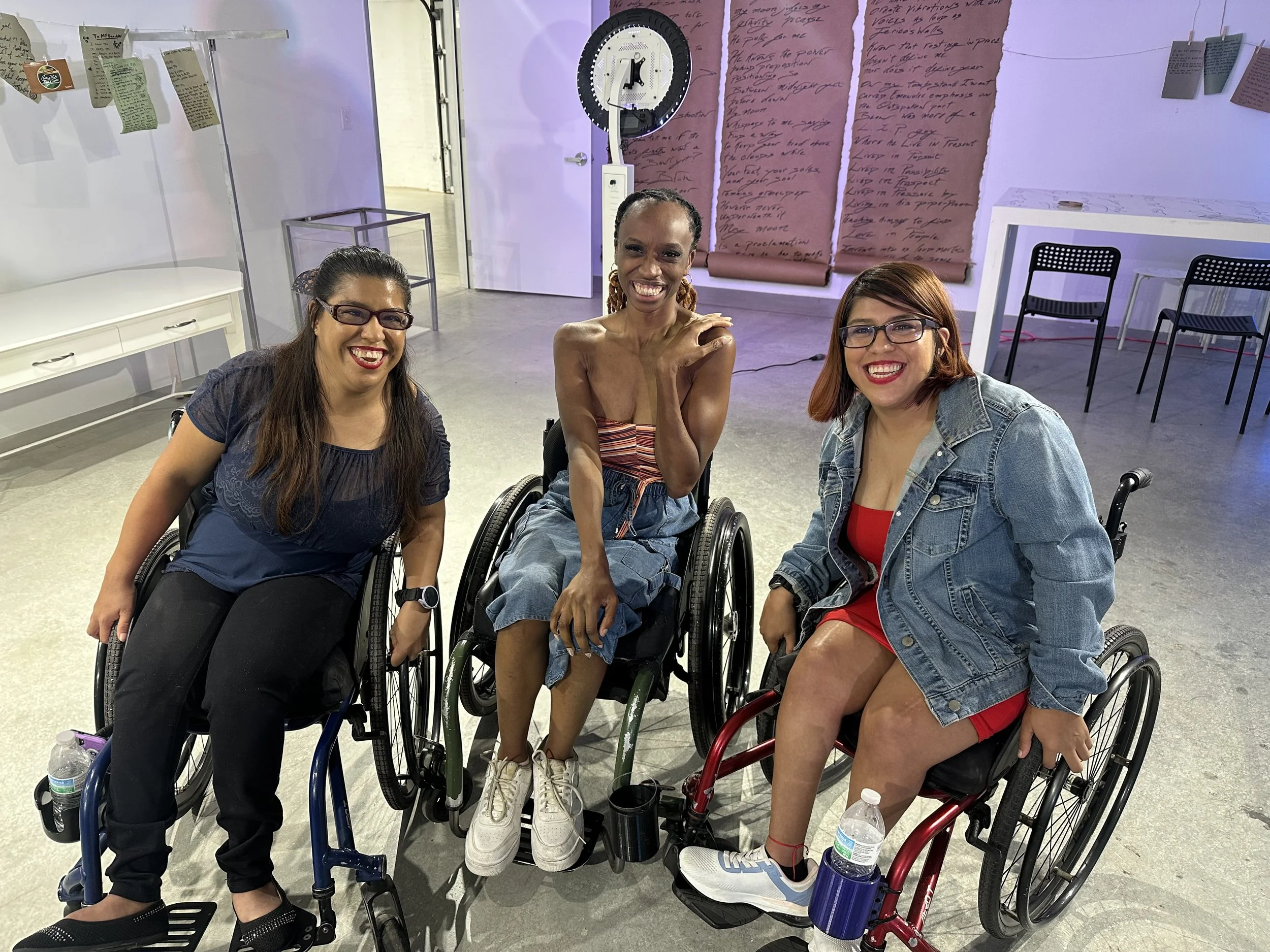 Three women in wheelchairs smiling and posing for the photo in an indoor space with purple lighting, with an art installation in the background.