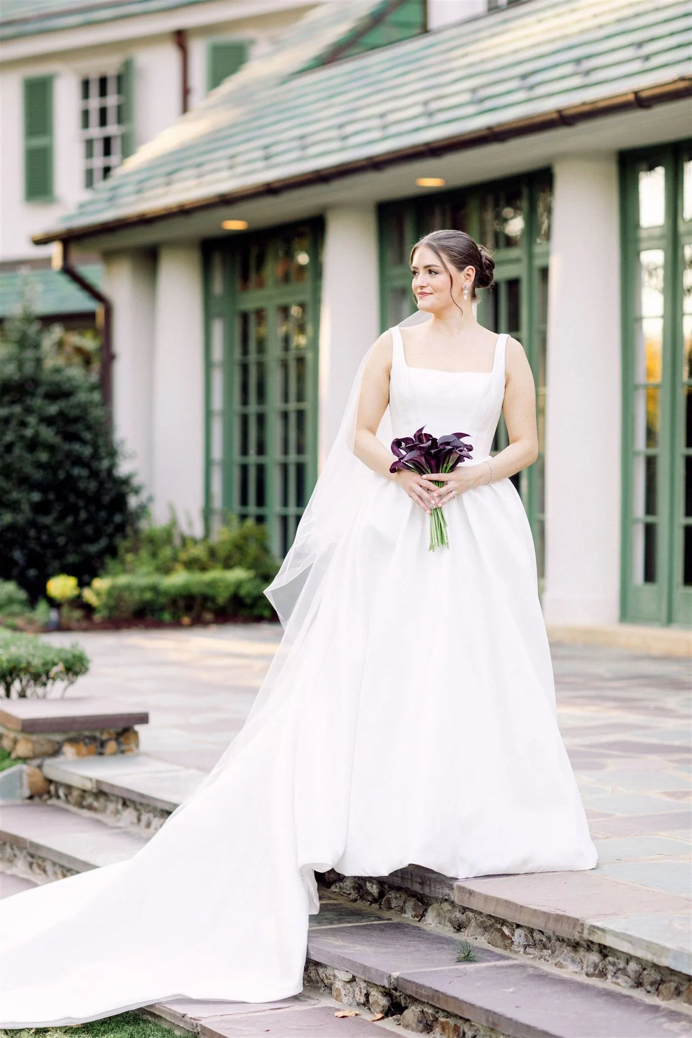 A bride in a white wedding gown holding dark purple calla lilies standing outside a building with large green windows and a stone walkway.