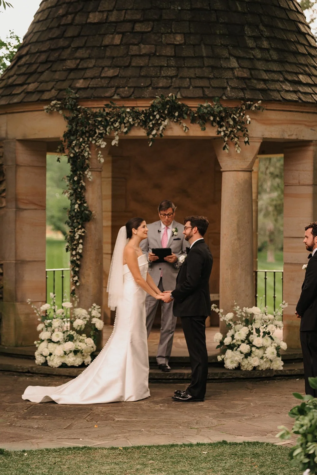 A bride and groom exchanging vows during an outdoor wedding ceremony in front of a wooden gazebo decorated with white flowers and greenery. The officiant stands behind them reading from a black book, with two groomsmen observing on either side.