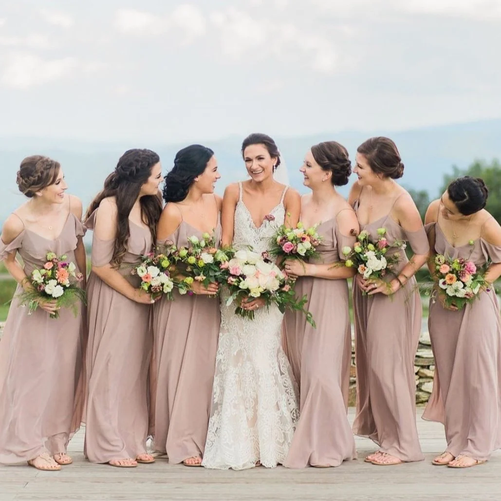 A bride and six bridesmaids standing together outdoors, holding bouquets, smiling and laughing. The bride wears a lace wedding gown, and the bridesmaids wear matching taupe dresses.