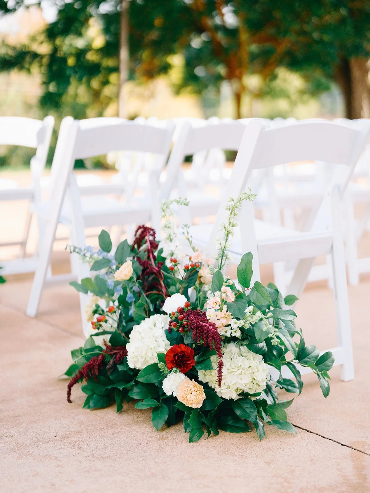 White folding chairs set up outdoors with a floral arrangement on the ground in front, featuring white, red, and pink flowers with green foliage, on a concrete surface with trees in the background.
