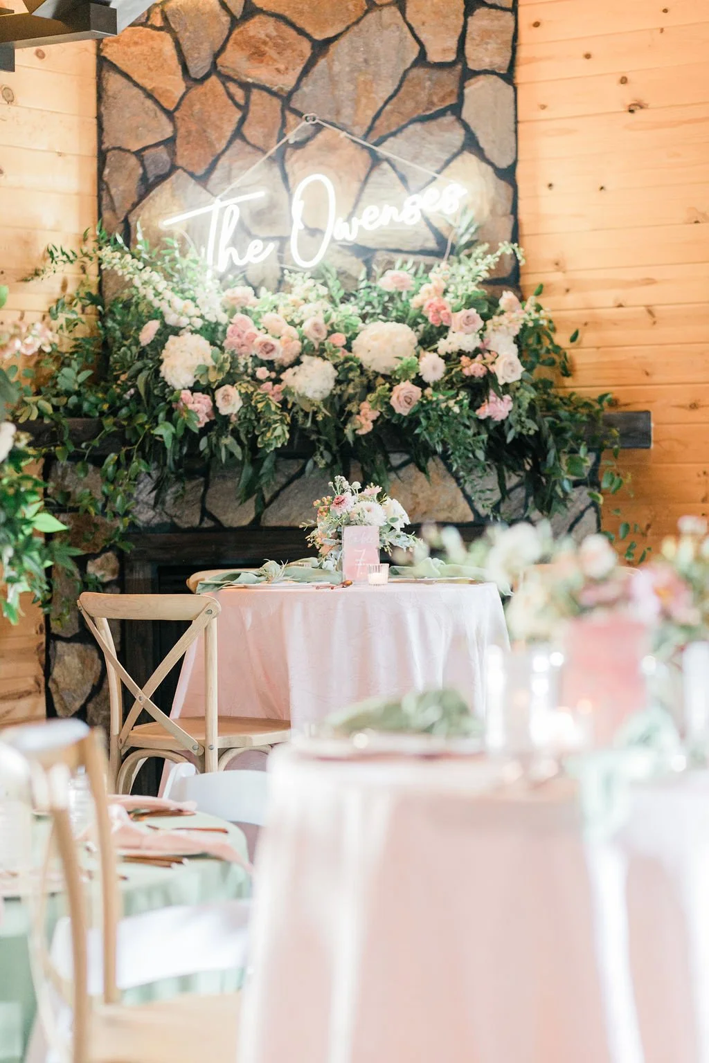 Decorated reception area with a stone fireplace, floral arrangements, and a neon sign that reads "The Ovenses" in cursive. Tables with pink and green tablecloths and chairs are in the foreground.