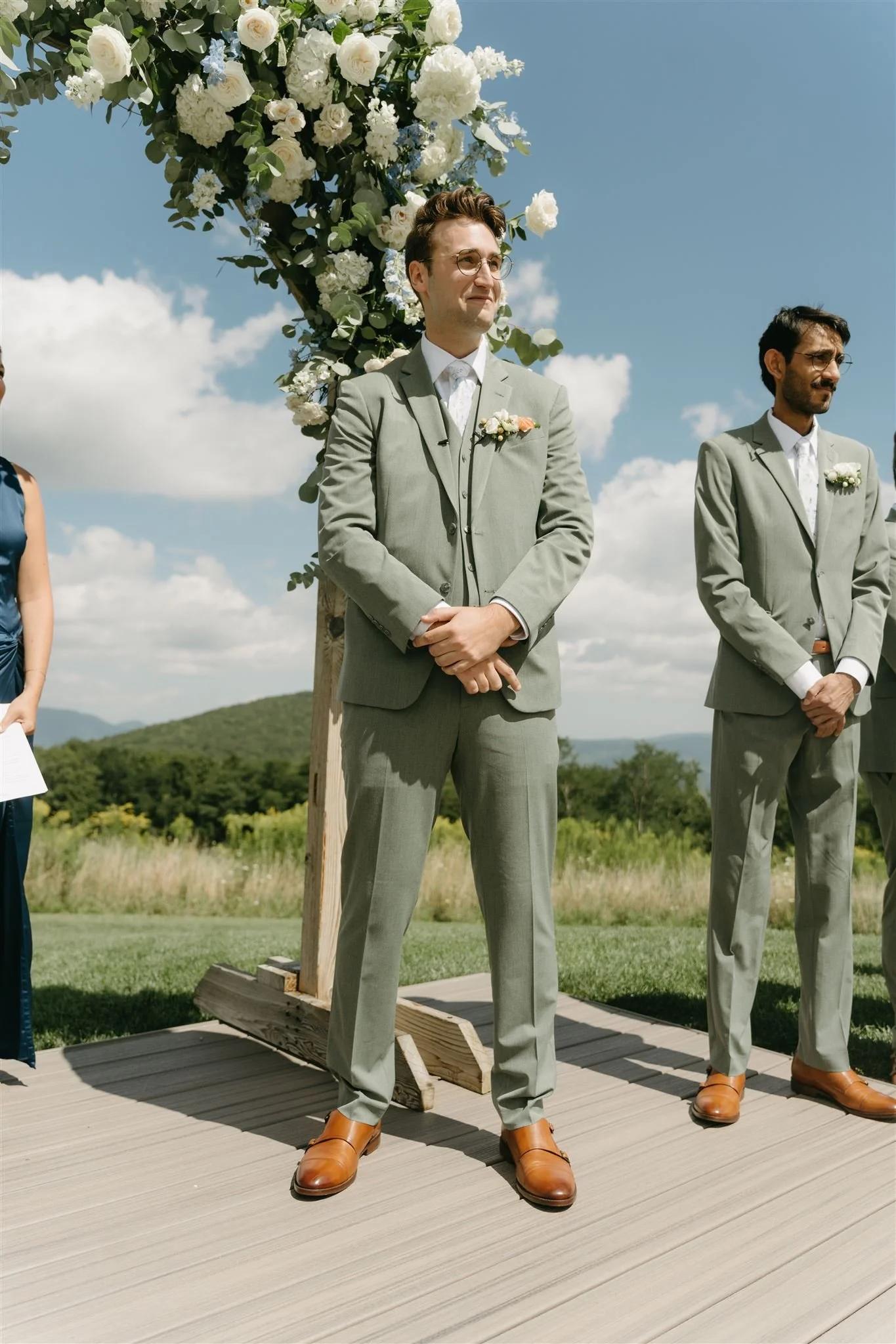 A groom dressed in a light gray suit with brown shoes, standing with hands clasped in front, during an outdoor wedding ceremony with a floral arch and blue sky background.