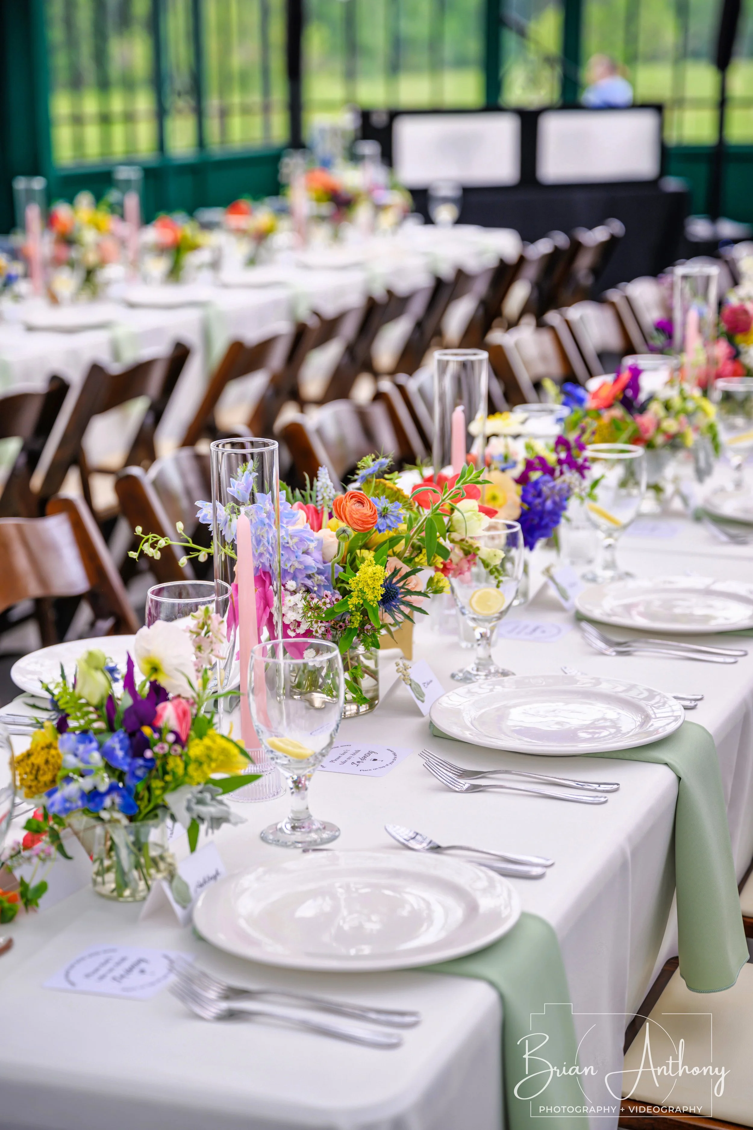 Elegant banquet table set with colorful floral centerpieces, white plates, silverware, pink candles, and glasses, ready for a wedding or celebration in a bright conservatory with green outdoor scenery.