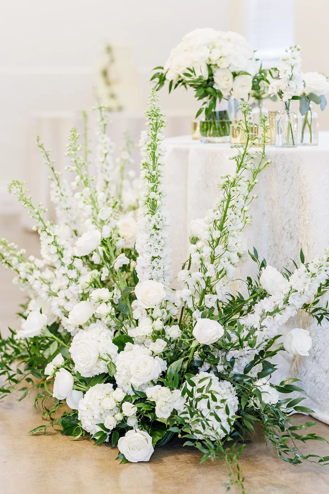 Elegant white floral arrangement with roses, hydrangeas, and greenery on a wooden floor, with a table of matching white flowers in glass vases in the background.