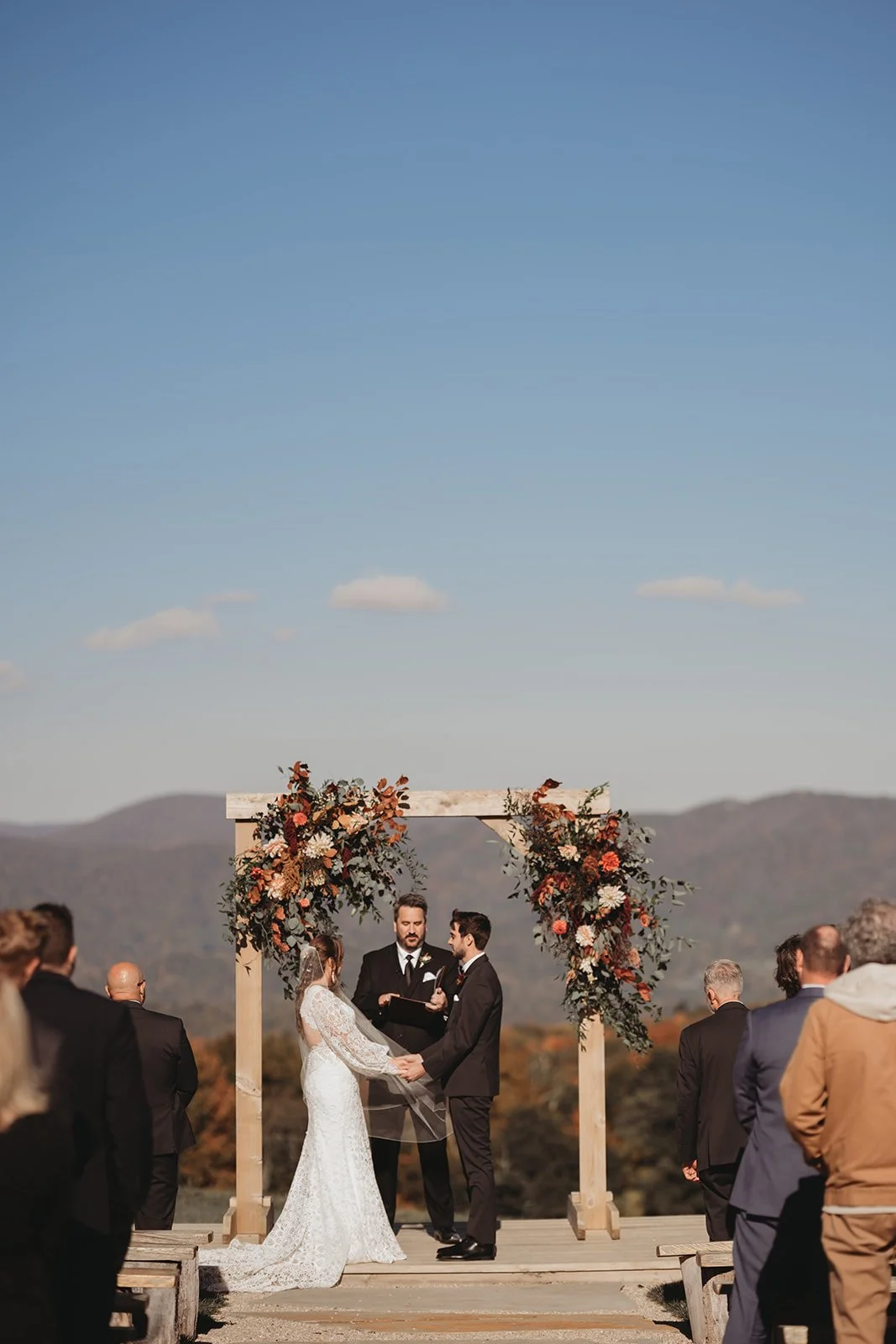 A couple getting married at an outdoor ceremony with guests standing around, a wooden arch decorated with flowers behind them, and mountains in the background.