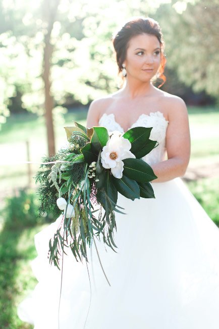 Bride in a white wedding dress holding a lush green and white bouquet outdoors.