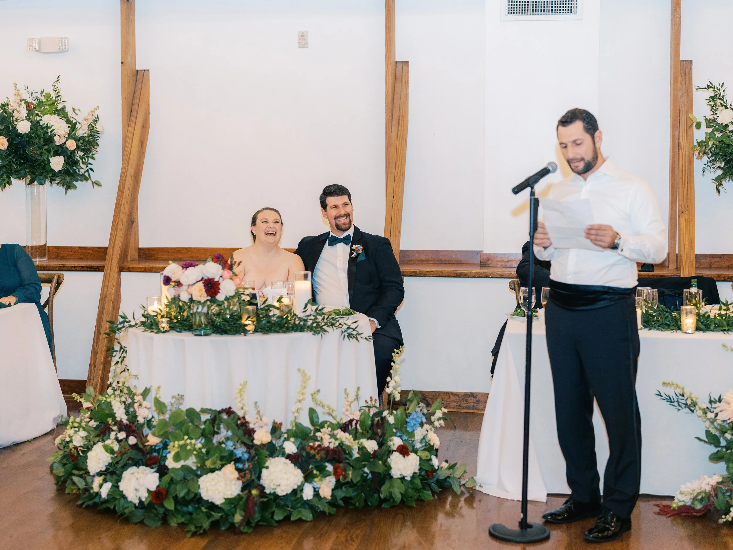 A man giving a speech at a wedding reception, standing at a microphone with a paper in hand, while the bride and groom seated at a decorated table, smiling and looking happy.