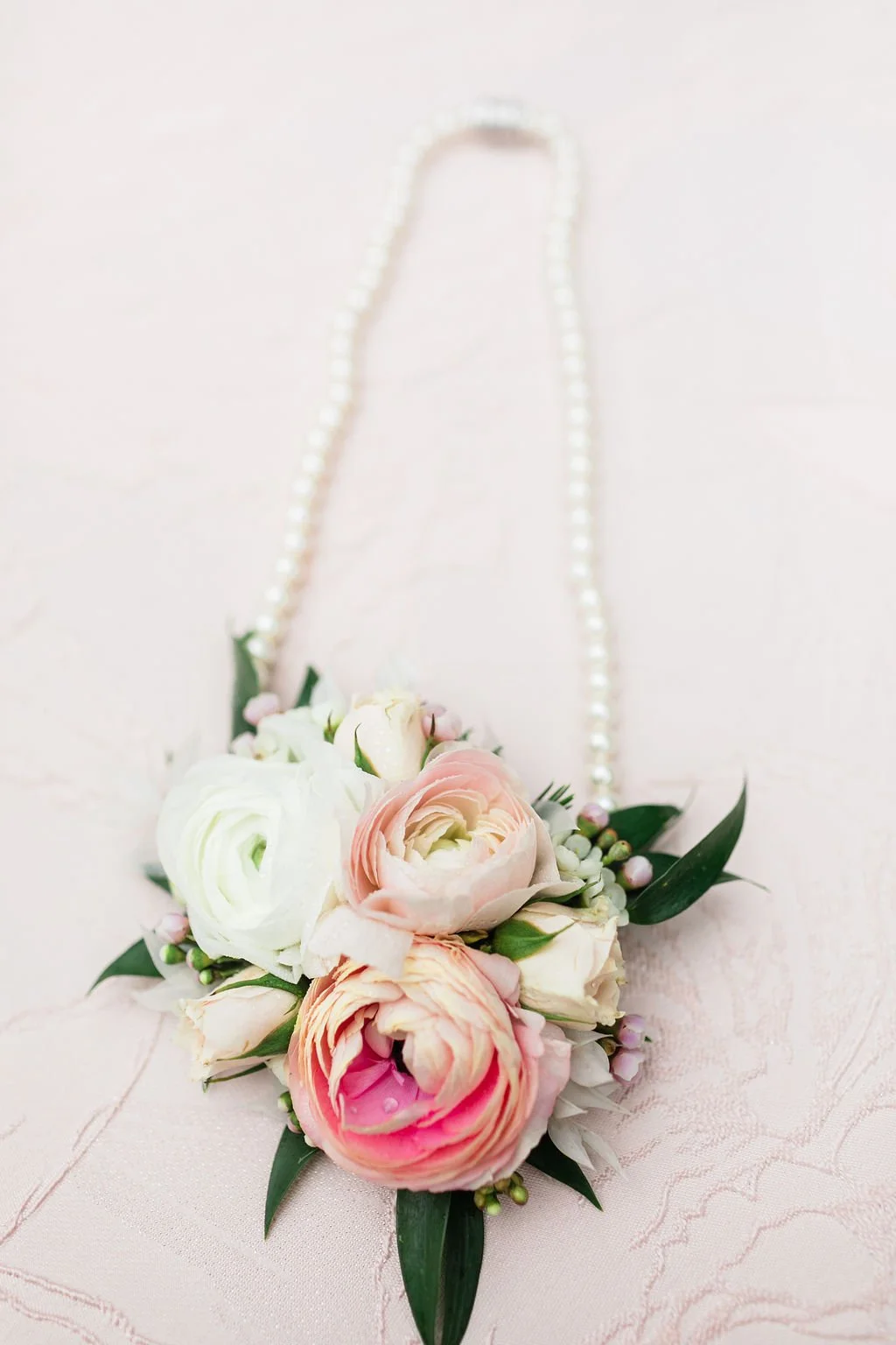 A floral bouquet with pink and white roses, white ranunculus, and green leaves, attached to a pearl necklace, resting on a light pink surface.