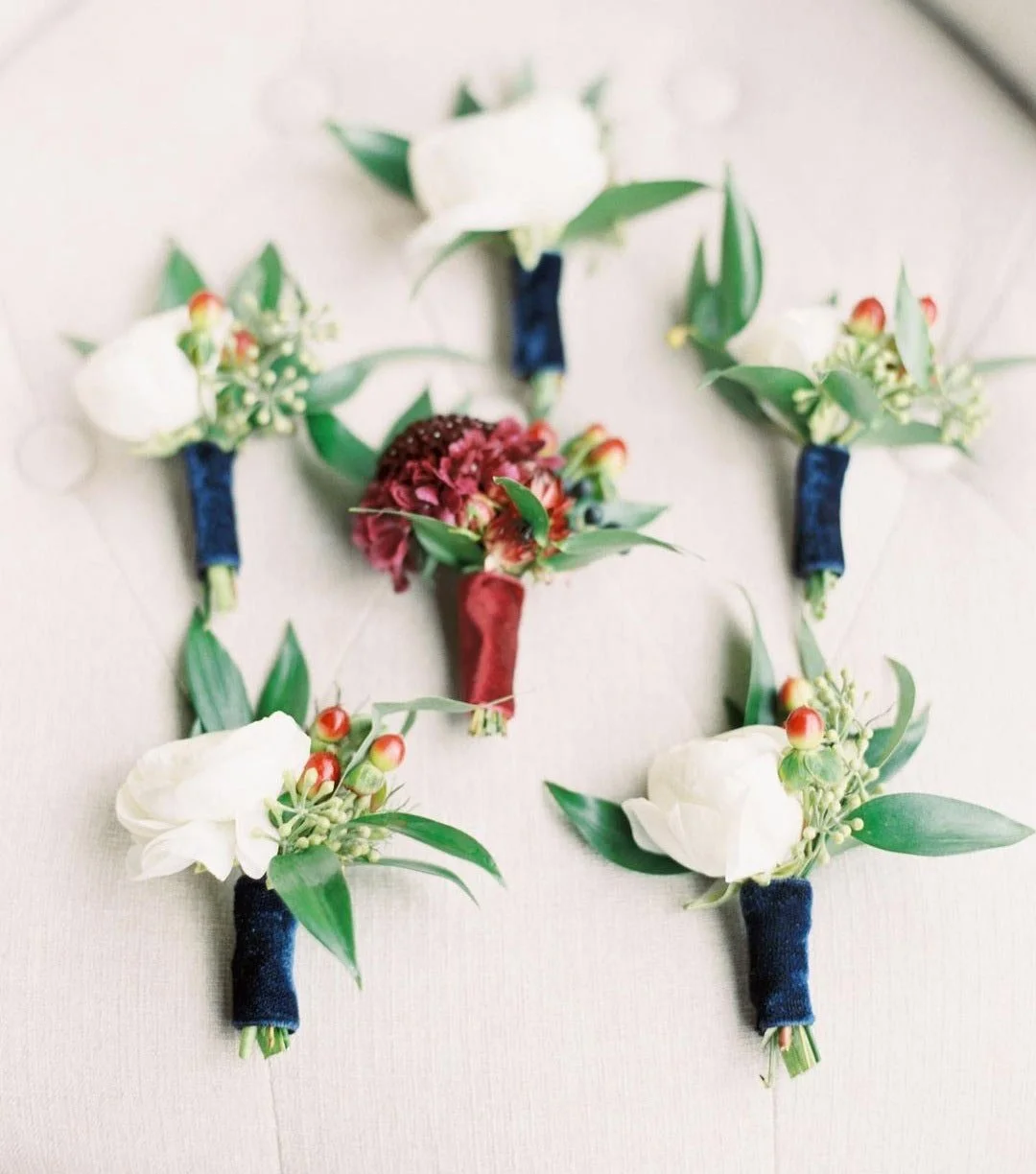 Multiple floral boutonnieres with white roses, greenery, and small red berries on a light fabric surface.