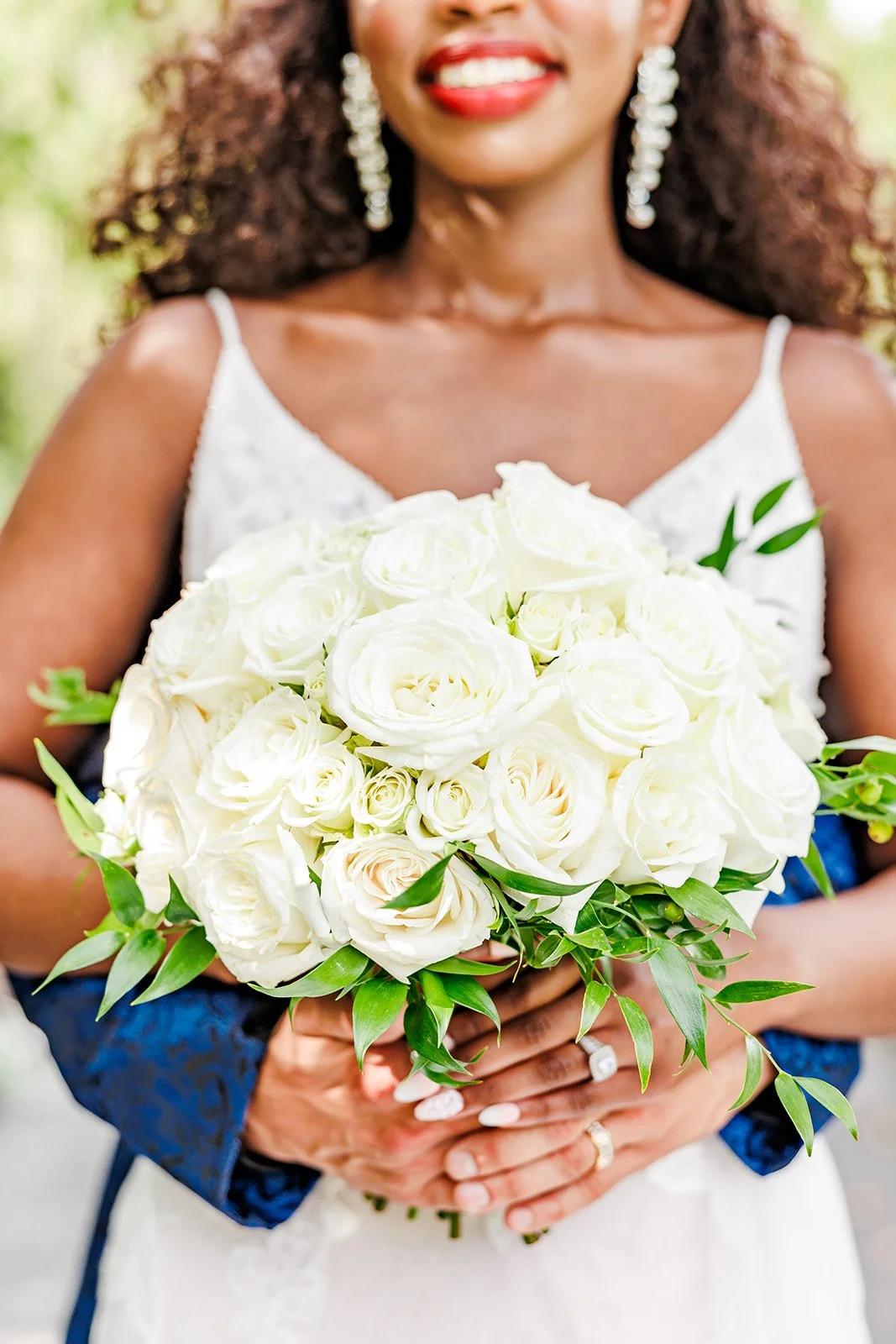 A woman in a white dress holding a bouquet of white roses and greenery, smiling with bright red lipstick and wearing dangling earrings.