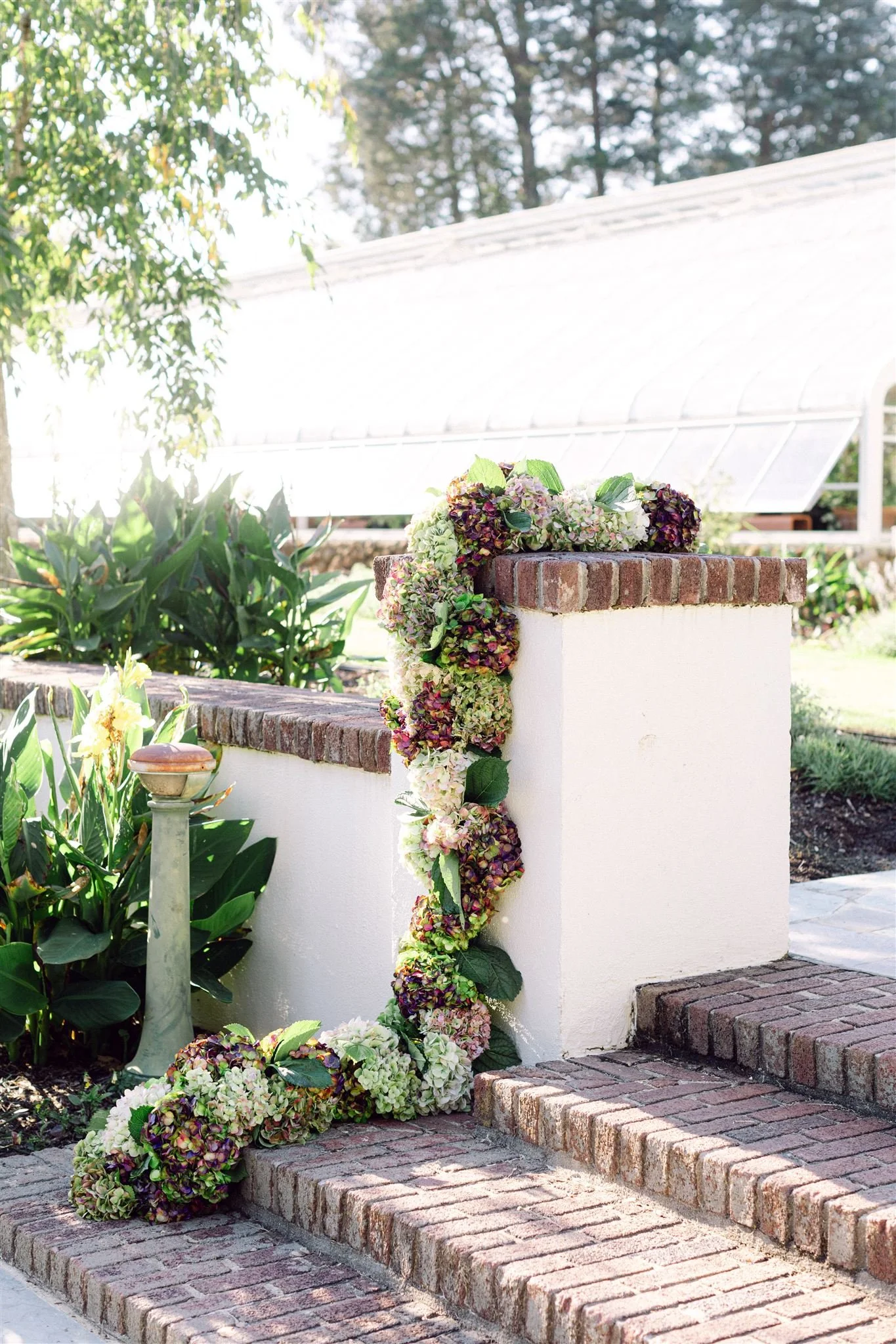 A floral arrangement of hydrangeas and other flowers in shades of purple, green, and white draped along a brick and white stucco wall outside in a garden setting.
