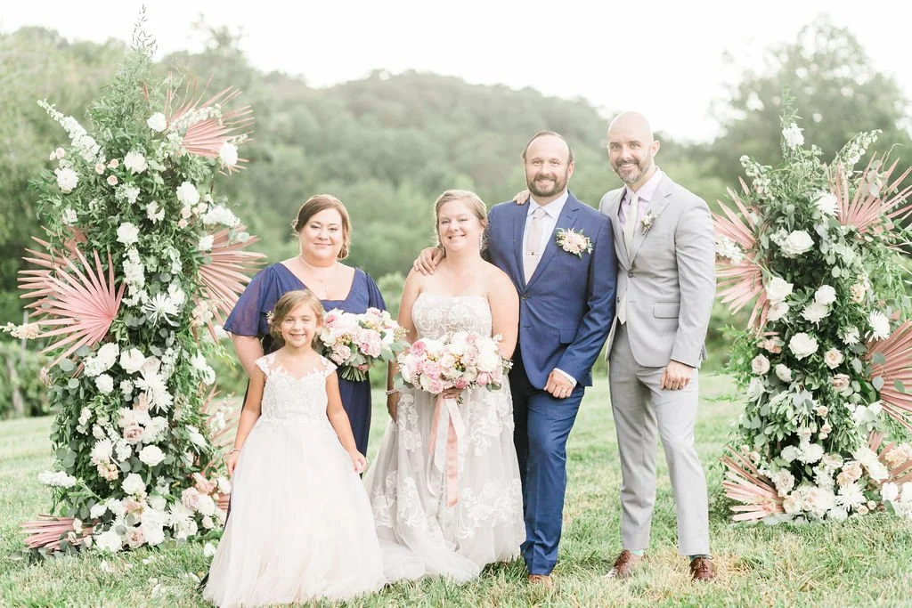 A wedding party standing outdoors under floral arches, including a bride and groom, a young girl, and three women dressed in formal attire, with green hills in the background.