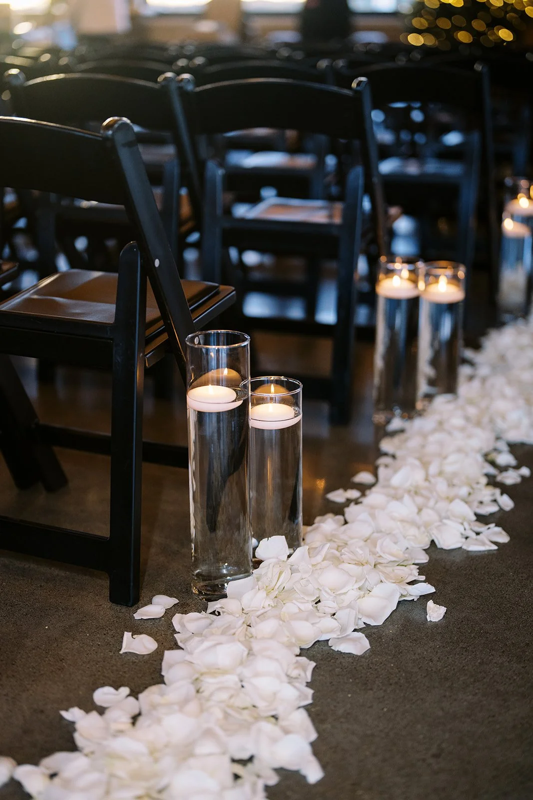 Candles in tall glass holders placed on the floor among white flower petals, with black chairs arranged in rows in the background, suggesting a wedding or romantic event setup.