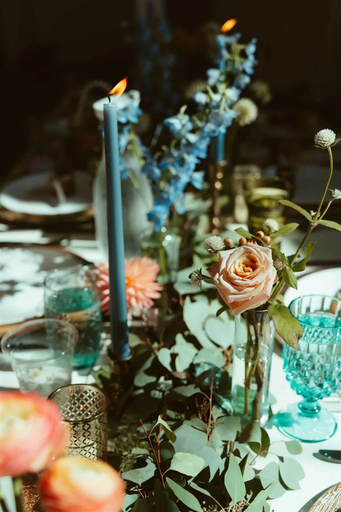 Vase with pink rose, blue delphiniums, and white flowers on a decorated table with glasses and candlesticks.